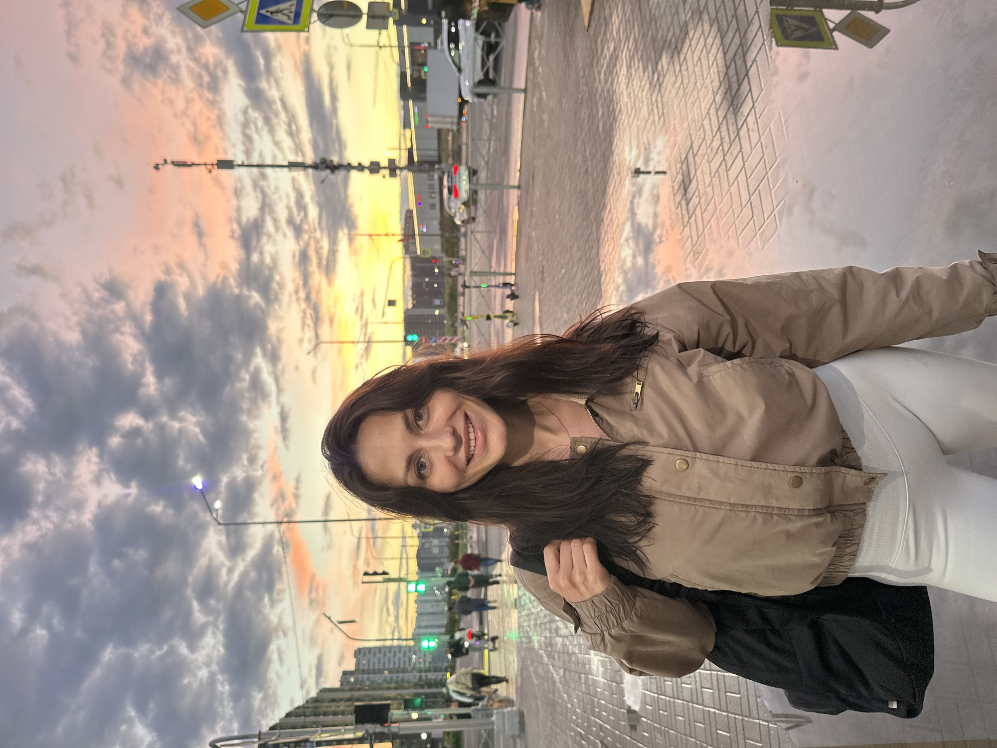 A woman with long, dark hair smiles at the camera, wearing a tan bomber jacket and white leggings. She has a black backpack slung over her left shoulder. The background depicts a city street at dusk, with a colorful sky transitioning from yellow to pink and grey clouds. Traffic lights are visible, showing green, and a few people and electric scooters are present on the sidewalk. Puddles on the paved ground reflect the sky and surrounding lights. In the distance, buildings and traffic are discernible.