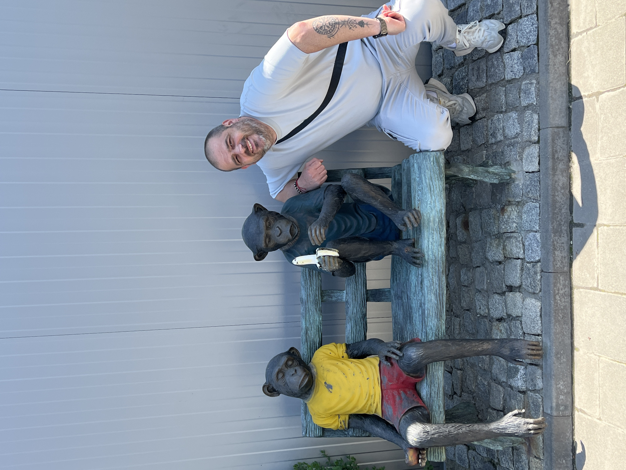 A man is posing with a bench of monkey statues. The man, with a beard and wearing a light-colored outfit, is kneeling next to the bench and smiling at the camera. The bench has two monkey statues, one lying down in a yellow shirt and red shorts, and the other sitting up and holding something in its hands. The photo appears to be taken in an outdoor setting, possibly Asenovgrad, Bulgaria, given the location provided. The background shows a wall and cobblestone pavement.