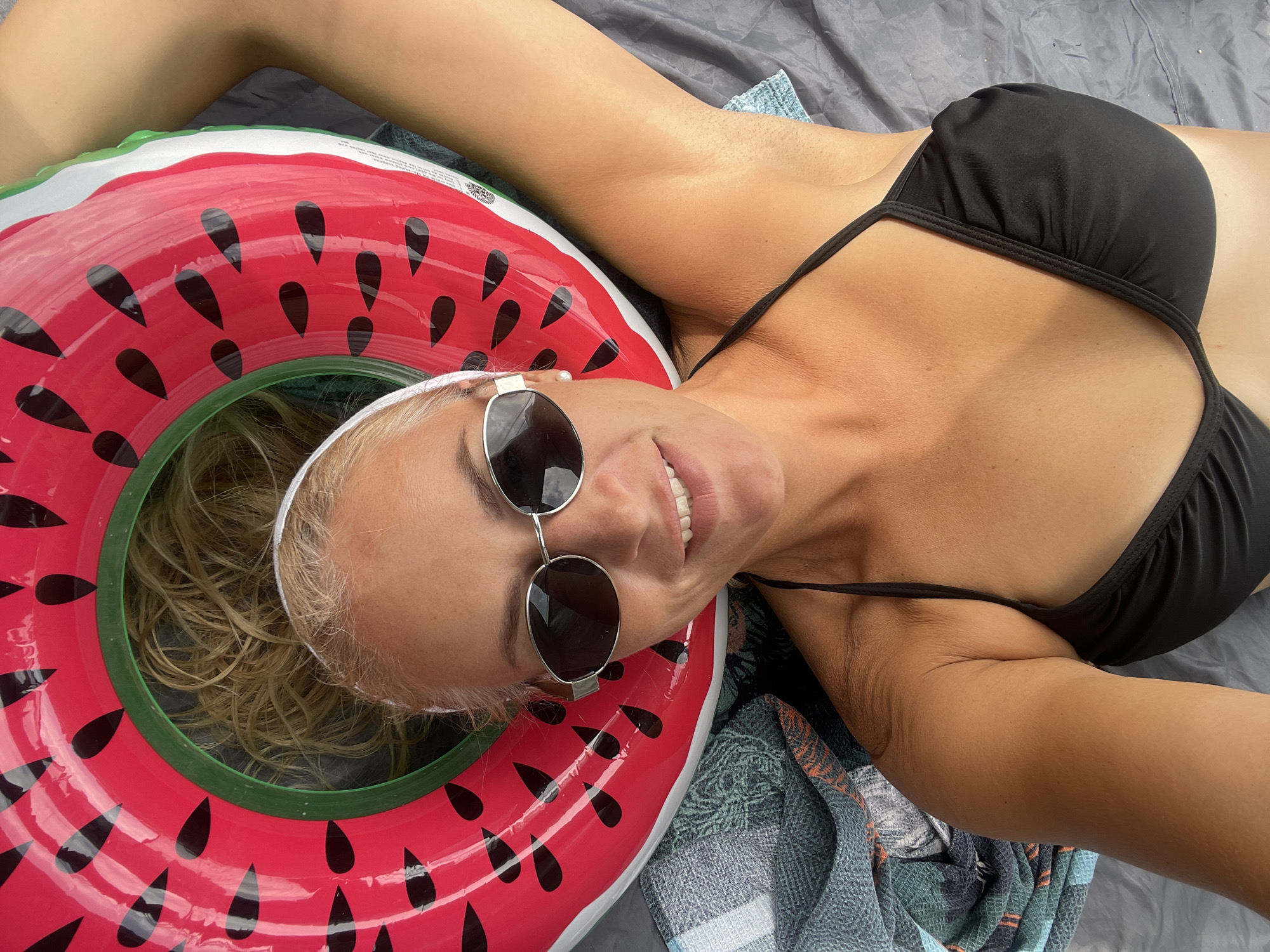 A woman is lying down, wearing a black bikini top and sunglasses, with a watermelon-shaped inflatable ring around her head. She is on a gray surface, possibly a beach towel or blanket, with another patterned towel to her left. Her hair is blonde and appears slightly messy. The lighting suggests it is daytime. The scene appears to be outdoors.
