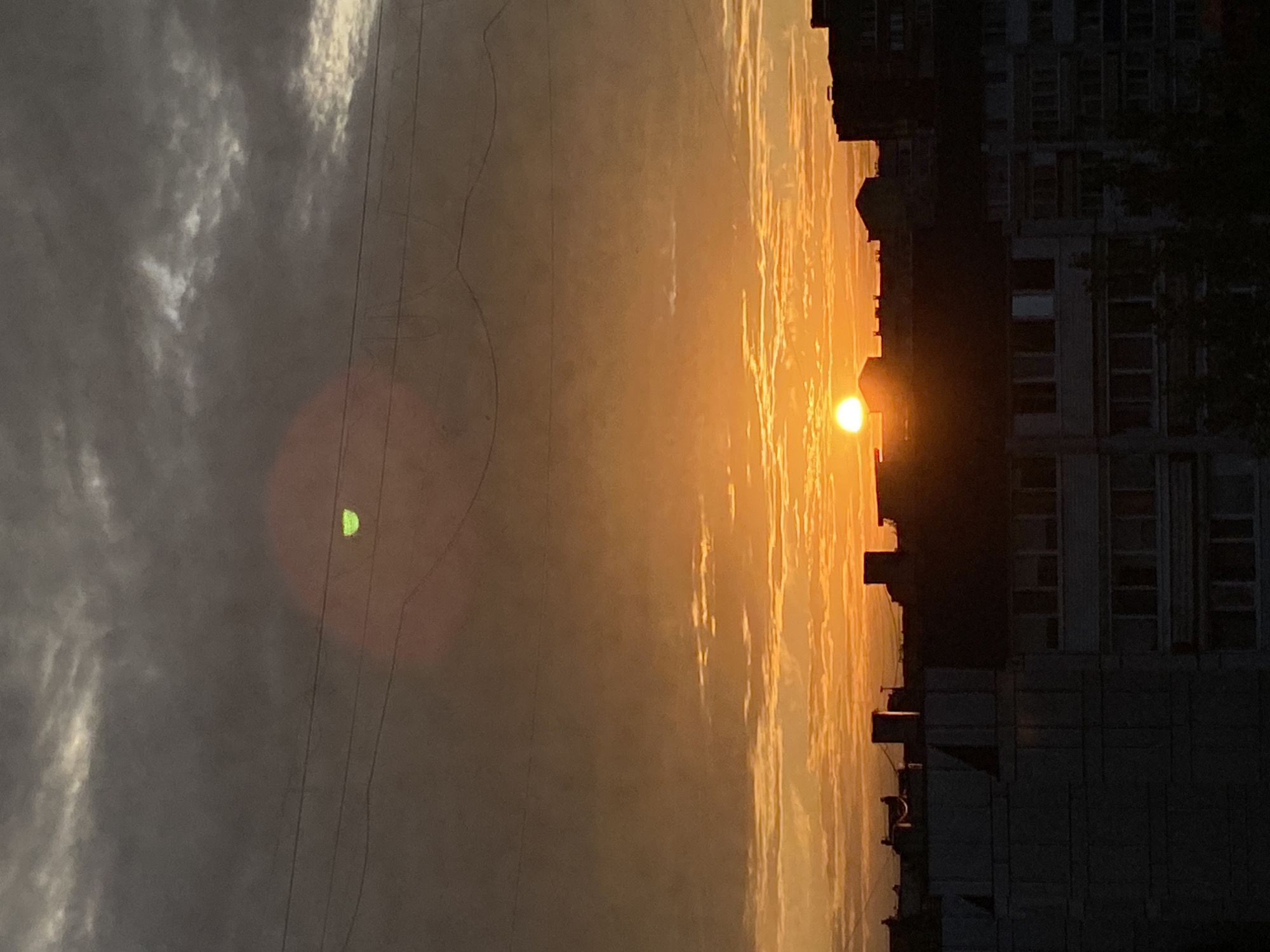 The image depicts a cityscape at sunset, likely in Chaikovsky, Russia, given the context provided. Silhouetted against a vibrant orange and yellow sky, several multi-story buildings form the skyline. The setting sun, a bright orb, is partially obscured by the buildings. Overhead, dark clouds are interspersed with streaks of lighter tones, suggesting a dramatic sky. Several power lines stretch across the upper portion of the frame. The lower part of the image is dominated by the dark silhouettes of apartment buildings, their windows appearing as dark rectangles. A faint, circular, green lens flare is visible in the upper portion of the sky, to the left of the sun. The scene is devoid of any visible people or animals.