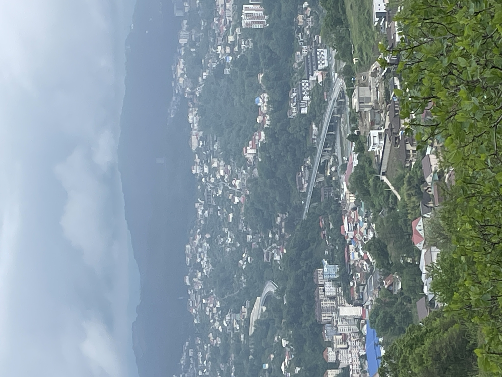 The image shows a panoramic view of a hillside city in Sochi, Russia, captured from an elevated vantage point. The scene is dominated by densely packed buildings and houses scattered across the green slopes. In the foreground, there are trees. A winding road or highway is visible, traversing the urban landscape. In the background, a hazy mountain range borders the horizon. The overall atmosphere suggests a cloudy day.