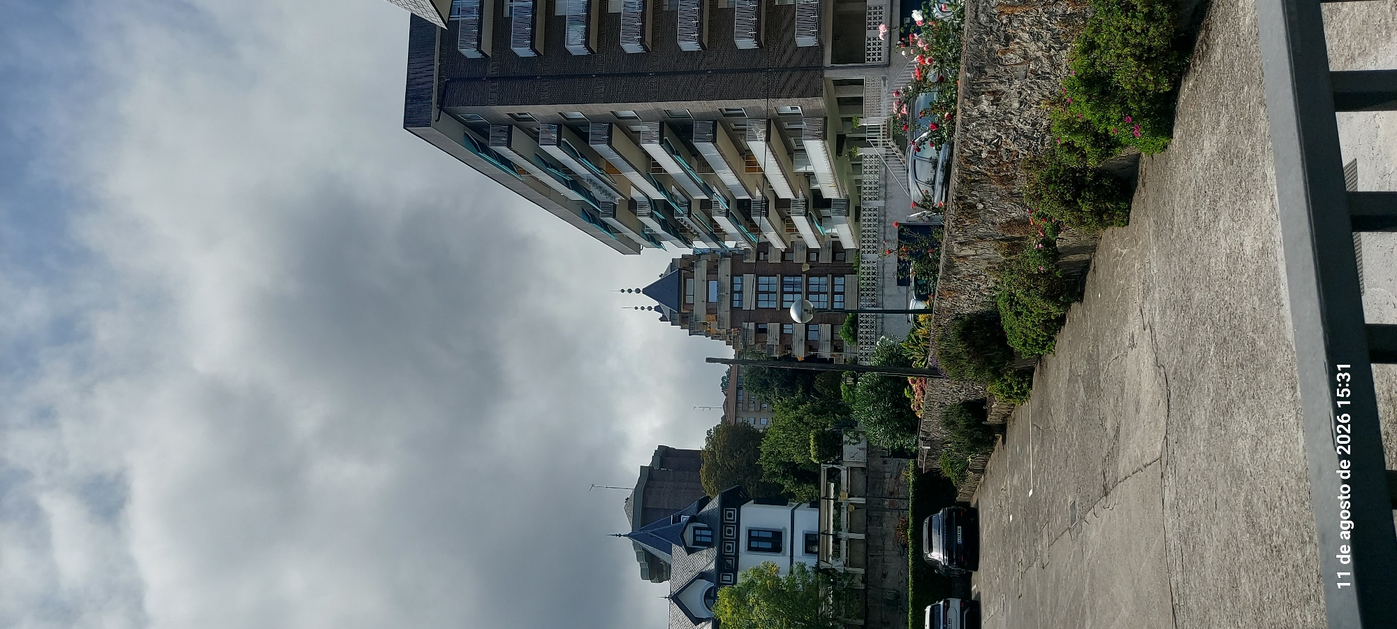 The image shows a street scene in Castro-Urdiales, Spain, on a cloudy day, as indicated by the date stamp "11 de agosto de 2026 15:31". In the foreground, a paved area with parking spaces is visible, bordered by a stone wall supporting a garden with lush green bushes and flowering plants, including roses. Several cars are parked in the spaces. Behind the wall, a street runs alongside apartment buildings.
One prominent building is a tall, multi-story apartment complex with balconies. To its left, another building with a distinctive tiered roof design and balconies is visible. Further to the left, a building with a dark, conical roof and a white facade with dark trim, possibly a residential home or a historic structure, is present, with a cylindrical tower behind it. The sky is overcast with dramatic grey clouds.