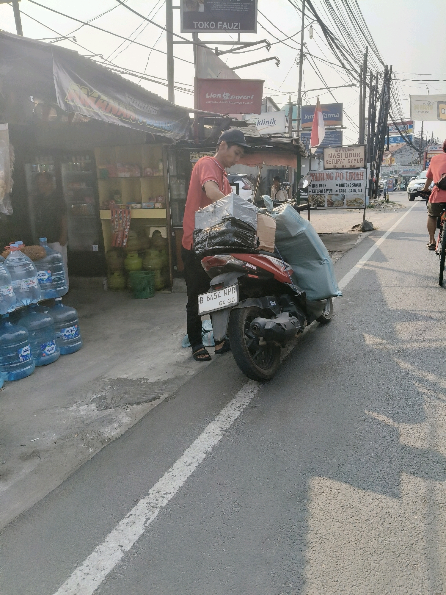 In South Tangerang, Indonesia, a man in a red shirt and black pants is standing next to a red and black motorcycle. The motorcycle is laden with packages wrapped in plastic and black bags, some covered with a light blue tarp. The man is wearing a black cap and appears to be in the process of loading or unloading the motorcycle. He is wearing sandals. To the left of the motorcycle, several large blue water jugs are stacked. Behind the man and motorcycle, a row of shops lines the street. One shop has a banner with Indonesian text. Gas cylinders are visible behind the shops. The street is paved with asphalt and marked with a white line. In the background, power lines crisscross the sky, and several signs for businesses, including "TOKO FAUZI," "Lion Parcel," "KIM FARMA," and "WARUNG NASI UDUK KETUPAT SAYUR IBU HALIMAH," are visible. A white car is driving on the street in the distance, and another person is riding a bicycle. The sun is casting shadows.