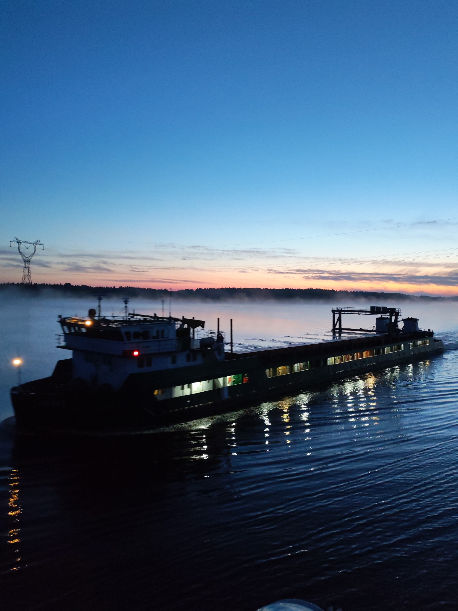 A barge travels on a body of water in Gari, Russia. The water is dark, with reflections of lights from the barge. The sky is blue, fading to orange near the horizon. Trees line the shore in the distance, obscured by fog. A power line tower stands to the left. The scene is illuminated by the barge's lights.