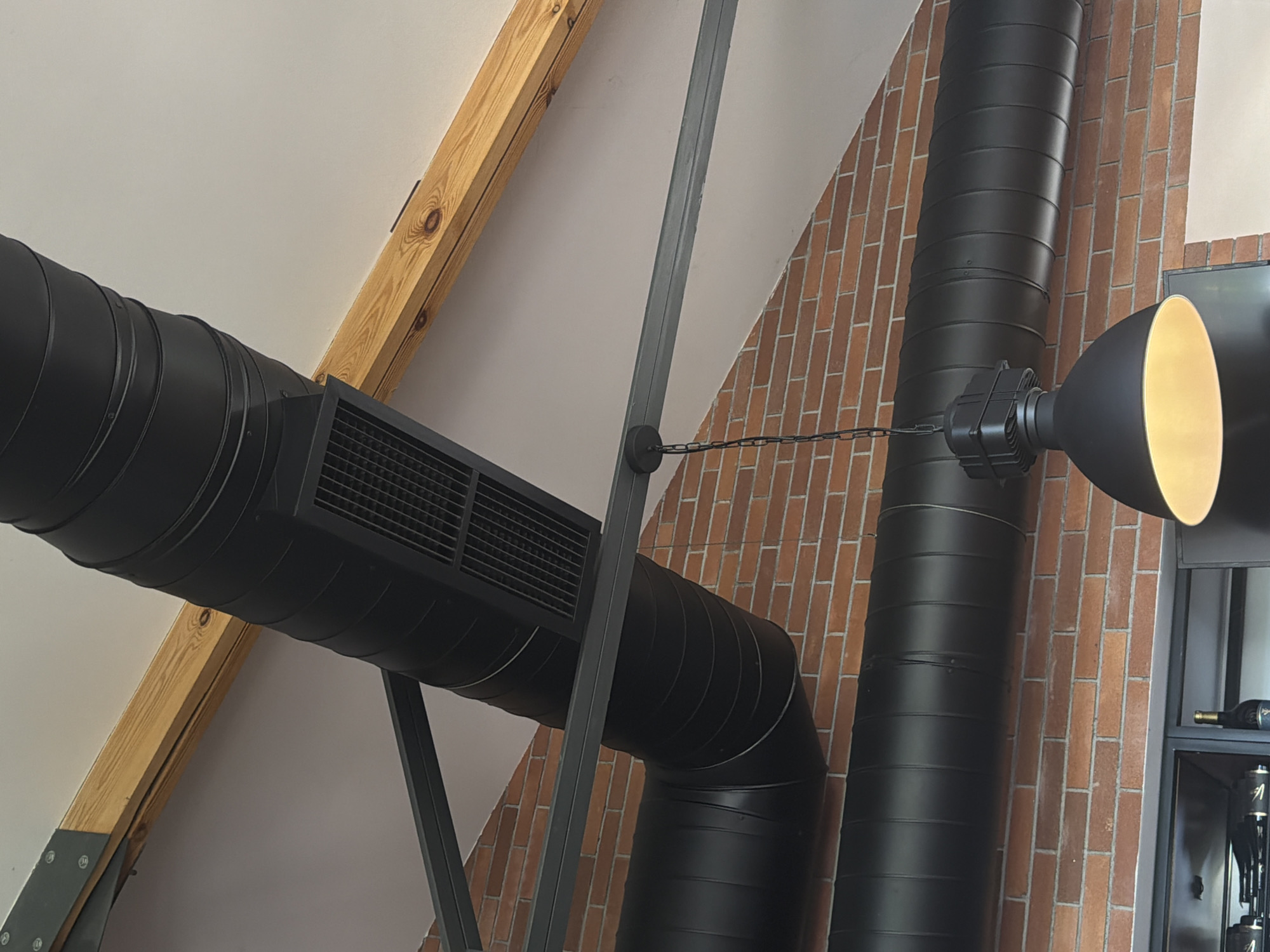 In Byurakan, Armenia, this image captures an interior view of a building's exposed ceiling. The ceiling features prominent black industrial-style ventilation ducts that run horizontally across the frame. These ducts are supported by dark gray metal beams, which are also attached to a light-colored pitched roof. A wooden beam, likely structural, runs diagonally across the upper portion of the image.
Visible in the lower part of the image are sections of exposed red brick wall. Hanging from one of the metal support beams is a black industrial pendant light with a wide, inverted bell-shaped shade that is illuminated from within. The overall aesthetic suggests a modern, industrial design, possibly in a restaurant, cafe, or open-plan office space. There are no people or animals visible in the image.