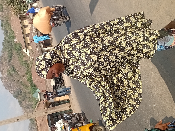 An elderly woman, clad in a black floral-patterned hijab and dress, occupies the foreground of a street scene. In the background, a sandy-colored mountain rises behind a row of modest buildings. Various people are visible: some stand by the buildings, while others ride motorcycles, one carrying a large, wrapped object. The scene is bathed in bright daylight.