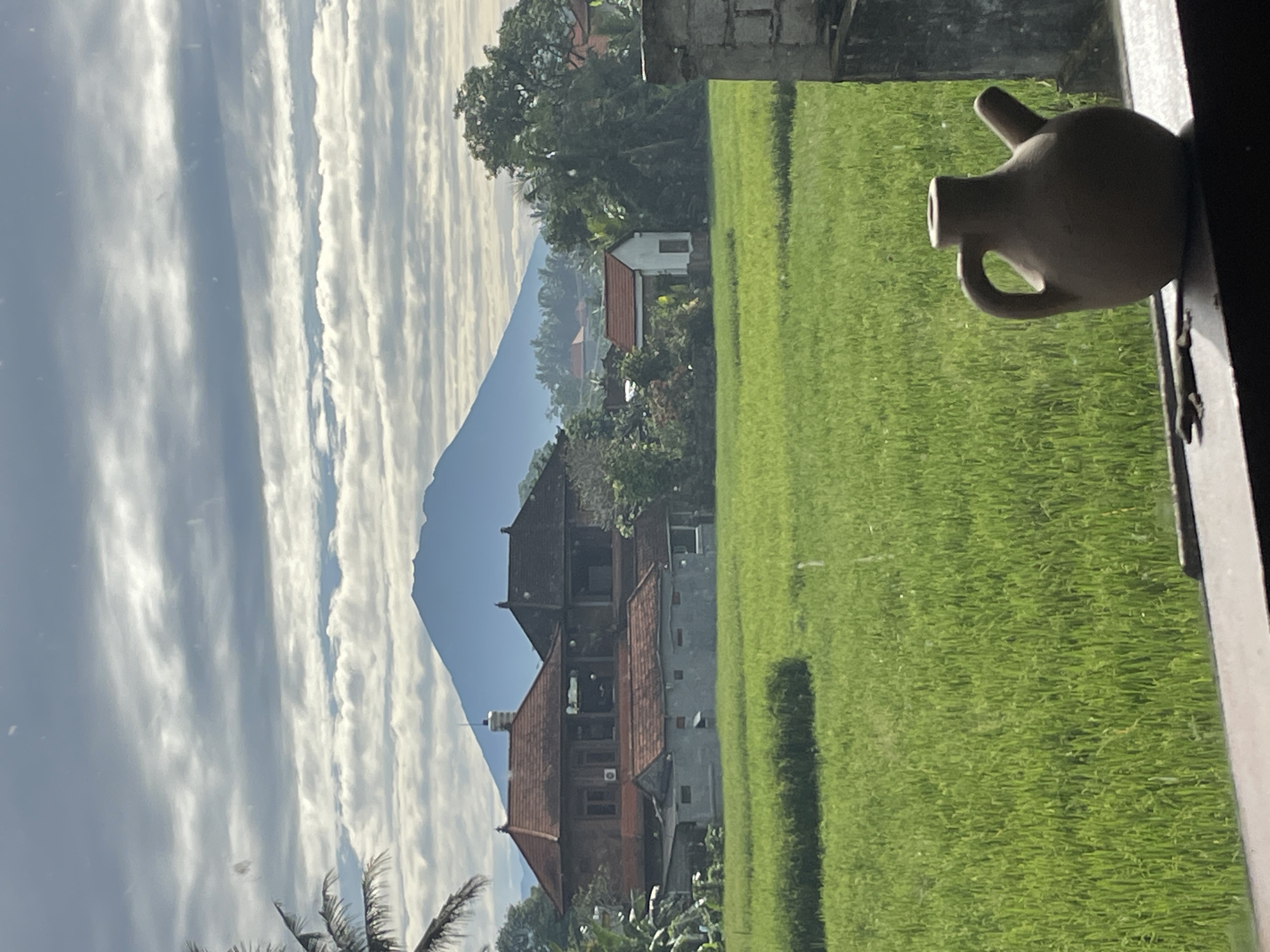 The image presents a tranquil, panoramic view of a rural landscape, likely observed from an interior space through a window. In the immediate foreground, on the right side, a small, light-colored ceramic jug with a handle and a spout rests on a windowsill, partially silhouetted.
The midground is dominated by a vast, lush green field, characteristic of rice paddies, extending horizontally across the frame. A darker, horizontal strip is visible within the field, possibly indicating a path or a different section of the crop. Scattered among rich green trees are several houses with traditional architecture, featuring distinct reddish-brown tiled roofs. One multi-storied house stands out with visible details, while a smaller, simpler white house is situated further back. Palm trees also dot the landscape.
In the background, a majestic mountain or volcano rises prominently, its peak clearly defined against the sky. The sky itself is a mix of clear blue and elongated, horizontal formations of white, wispy clouds, suggesting a bright but possibly partly cloudy day. The soft lighting indicates daytime, perhaps morning or late afternoon. The scene evokes a sense of peaceful natural beauty and rural life, typical of regions with rice cultivation and mountainous terrain. No text is visible in the image.