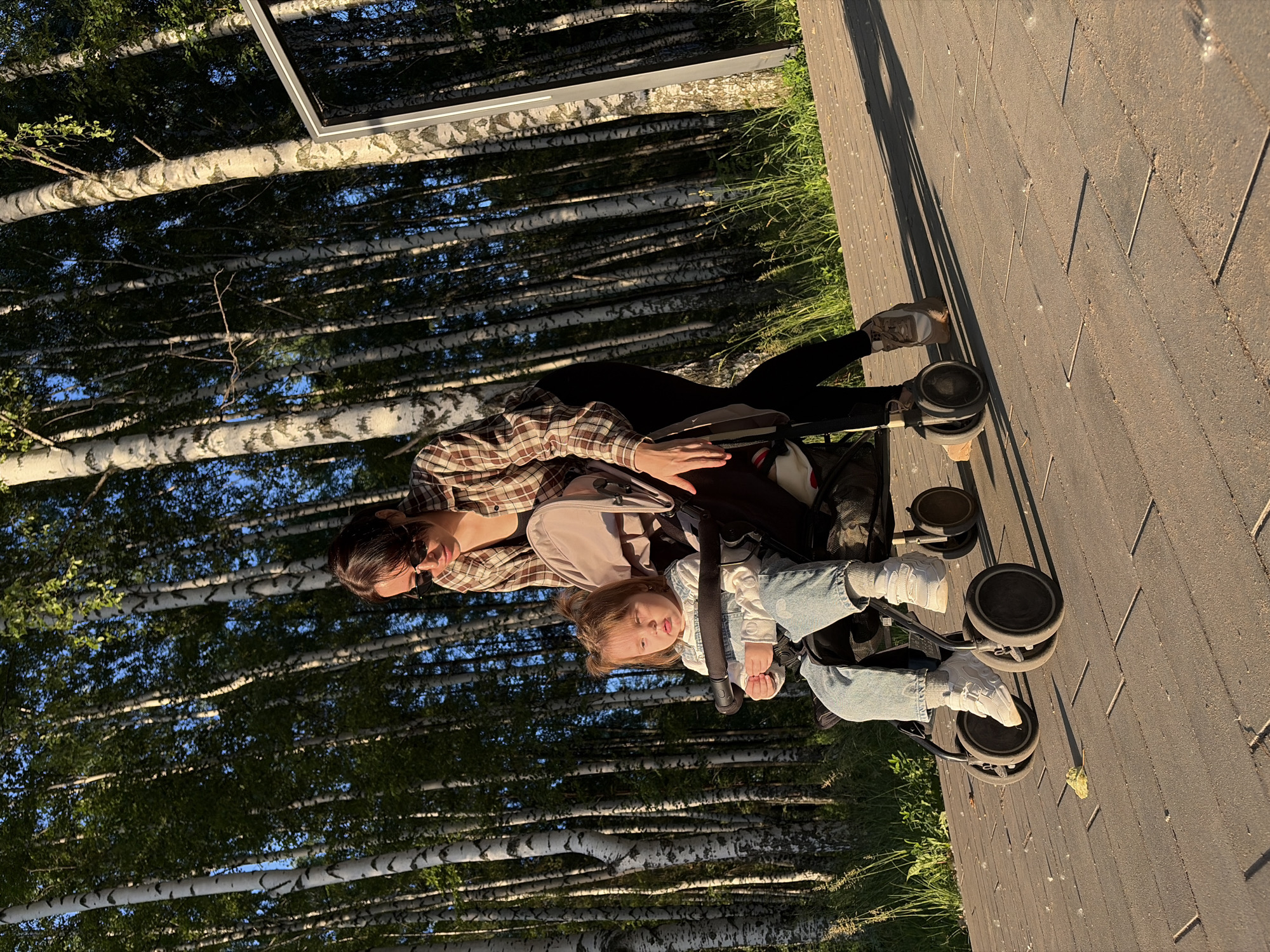 In Chaikovsky, Russia, a young child sits in a stroller on a paved walkway lined with birch trees. The child, wearing denim overalls and a long-sleeved shirt, looks to their left with a neutral expression. Behind the stroller, a woman in sunglasses and a plaid shirt pushes it. Her hands are on the stroller's handle, and she appears to be walking. The sun casts long shadows from the trees onto the walkway, indicating it is late afternoon or early evening. The background is dominated by the dense birch forest, with a modern white structure visible to the right. The lighting suggests a warm, sunny day.