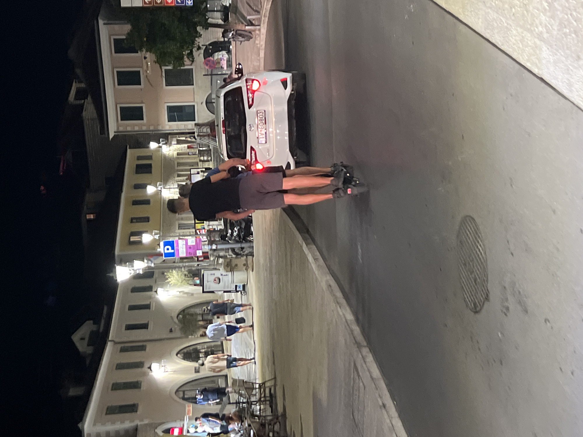 The image depicts a nighttime scene in the city of Herceg Novi, Montenegro. In the foreground, two individuals are riding on a single electric scooter on a paved road. The person in front is standing upright, while the person behind them is straddling them. Both are wearing casual clothing. Behind the scooter, a white Toyota Yaris hatchback is visible, its taillights illuminated. The license plate of the car reads "TV-BD803".
The background reveals a pedestrianized area lined with buildings. These structures appear to be commercial establishments, possibly shops or restaurants, with arched entrances and lit windows. Streetlights cast a warm glow on the scene, illuminating the pavement and the facades of the buildings. Several other people are walking in the background, and outdoor seating areas for cafes or restaurants are present. The overall atmosphere suggests a lively evening in a European coastal town. The presence of the scooter on the road and the pedestrian area indicates a mixed-use urban environment.