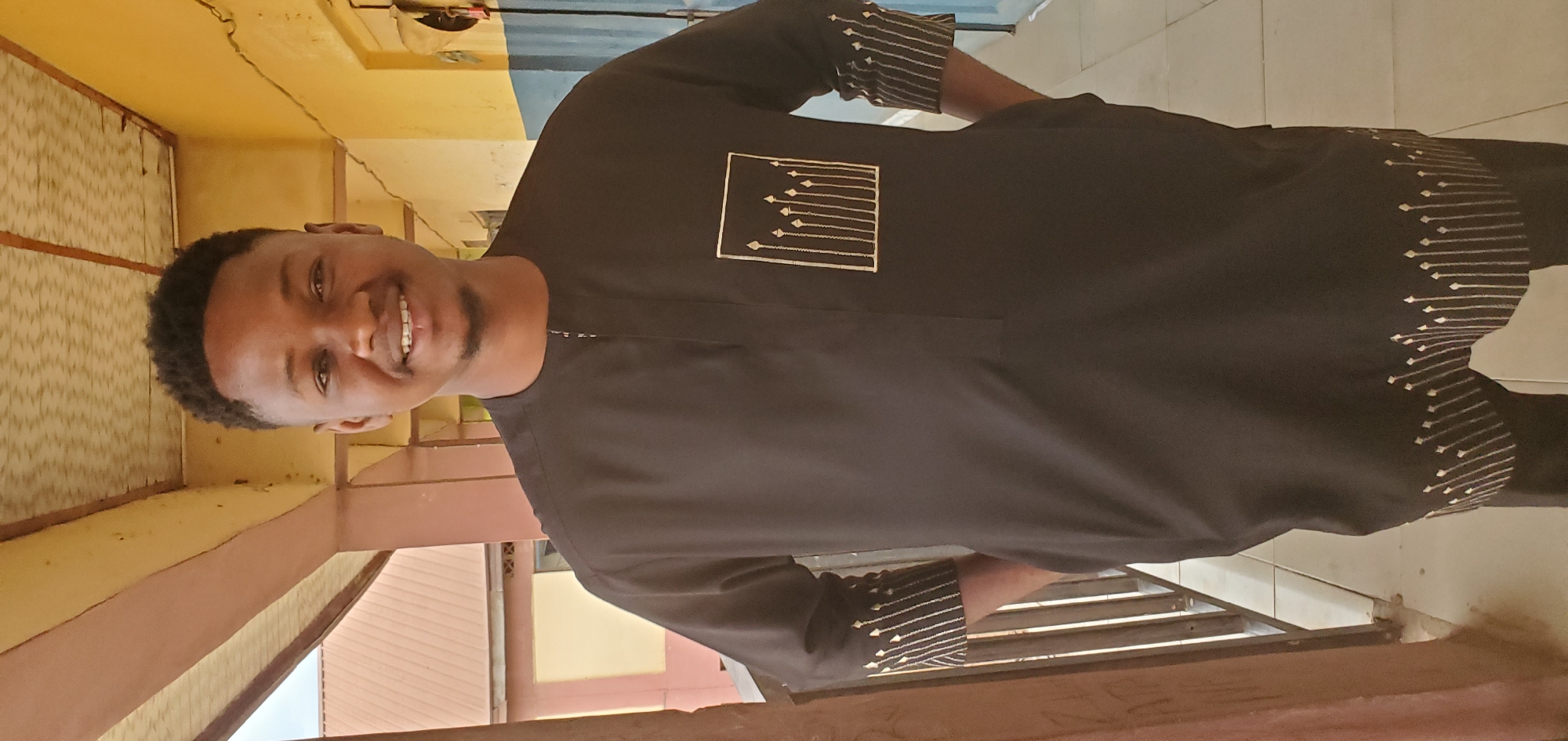 A smiling man with short, dark hair and a beard stands in front of a yellow and brown building. He is wearing a black, long-sleeved shirt with white designs on the chest, sleeves, and hem. The location is in Mararaba, Nigeria. The environment includes a building with a railing and a partly visible sky.