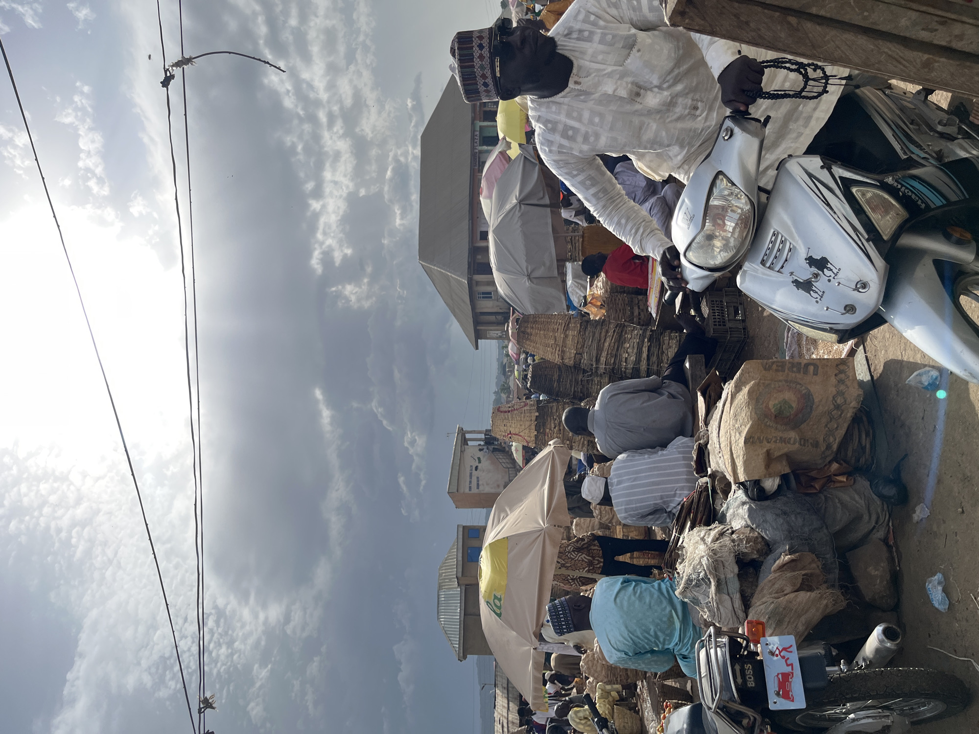 This is a medium shot of a busy marketplace in Jos, Nigeria, likely on a cloudy day with intermittent sunshine. In the foreground, a man wearing a white patterned tunic and a patterned cap is riding a silver scooter. He is wearing sunglasses and holding a set of prayer beads in his right hand. The scooter has decals of two polo players on its front. Behind the scooter, there are various goods piled up, including sacks and baskets. To the left of the scooter, the rear of another motorcycle is visible, with a license plate that reads "BOSS" and features a cartoon drawing of a car and people.
In the middle ground, several people are seated or standing, many of them dressed in traditional attire. Some are under large umbrellas, one of which is yellow and branded with green lettering. Woven baskets and other market goods are stacked high in the background. In the distance, buildings with corrugated metal roofs are visible. The sky is filled with dramatic clouds, with patches of bright sunlight breaking through, creating strong contrasts and casting shadows on the market. The overall impression is one of a vibrant, active, and somewhat chaotic marketplace.