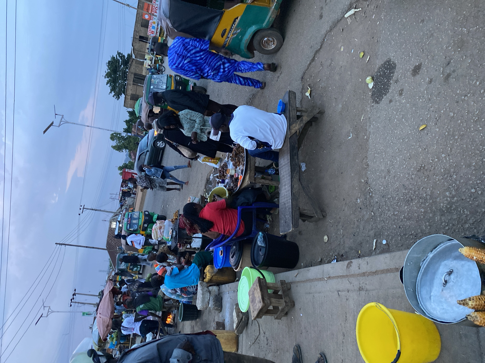 The image depicts a bustling street scene in Bukuru, Nigeria, likely a market area. In the foreground, a person wearing a blue and white patterned garment is walking away from the camera towards a green and yellow auto-rickshaw. Nearby, another individual in a white t-shirt and blue jeans sits on a wooden bench. To the left, a large yellow bucket sits on the ground next to a metal pot with corn on the cob. Various other people are visible in the mid-ground, some standing and others seated, engaged in activities that suggest commerce or casual interaction. Several auto-rickshaws, common modes of transportation in Nigeria, are parked or in motion. The environment appears to be a dusty, paved street lined with makeshift stalls and structures, indicating a lively outdoor market or roadside gathering. The sky is overcast, suggesting either early morning or late afternoon.