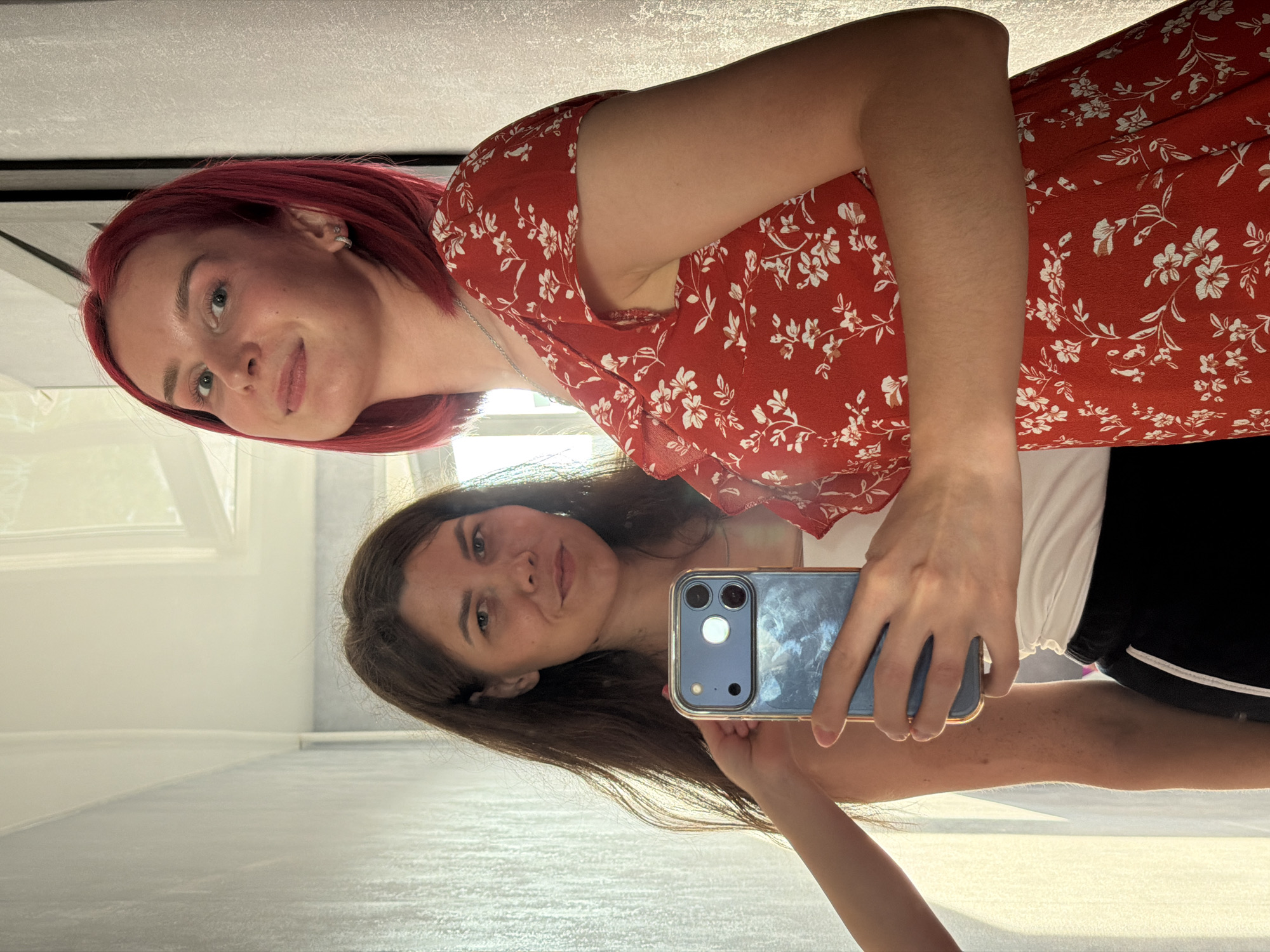 Two women are reflected in a mirror, taking a selfie. The woman on the right has short, bright red hair and is wearing a red floral dress. The woman on the left has long, dark brown hair and is wearing a white tank top and black athletic pants. A hand holding a phone is visible in the foreground, obscuring part of the woman on the left's face and body. The background appears to be a brightly lit interior space with light-colored walls and what looks like a window or doorway. There are no visible elements that specifically indicate the location is Chaikovsky, Russia.