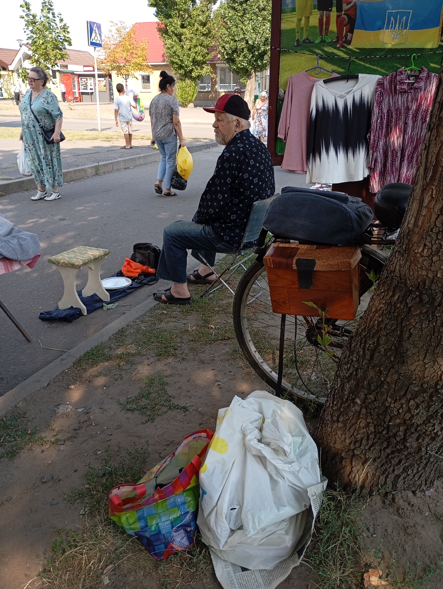 In the city of Horishni Plavni, Ukraine, on a sunny day, an outdoor market scene unfolds. A man in a patterned shirt and a red cap sits on a folding chair, facing away from the camera. Beside him, a bicycle is parked, fitted with a wooden box on the rear rack. On the ground near the bicycle are various items, including a backpack and a white plastic bag.
In the background, several individuals are visible. A woman in a light blue floral dress walks away from the viewer, carrying bags. Another woman in jeans and a patterned top walks towards the background, also carrying shopping bags. Further back, a young boy holds a colorful inflatable toy. The market appears to be set up along a sidewalk, with buildings and trees lining the street. In the distance, a Ukrainian flag is displayed. The ground is a mix of pavement and dirt, with some patches of grass.
