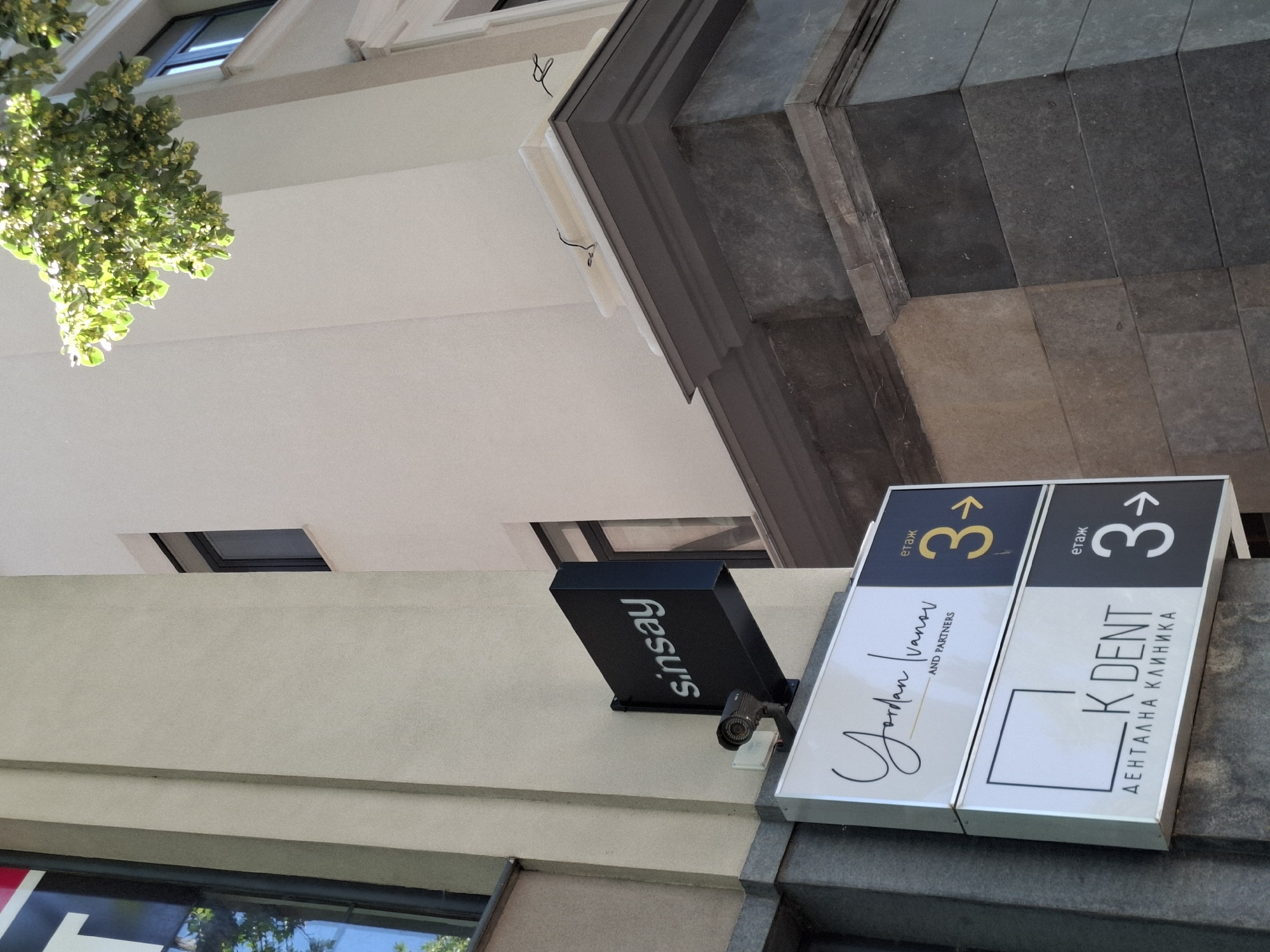 The image shows a modern building exterior with signs indicating businesses on the third floor. A black sign with white lettering reads "sinsay" above two stacked illuminated signs. The upper sign displays "Jordan Ivanov AND PARTNERS" in stylized script and sans-serif fonts, with a yellow "3" indicating the floor. The lower sign features a square logo and "K DENT" in sans-serif font, with "ДЕНТАЛНА КЛИНИКА" below it. This sign also shows a white "3" indicating the floor. A security camera is mounted below the "sinsay" sign. The building's facade is a light-colored stucco, with dark stone cladding visible at the base of the signage. Lush green tree branches with small yellow flowers overhang the top right of the image. The lighting suggests it is daytime, with sunlight casting shadows.