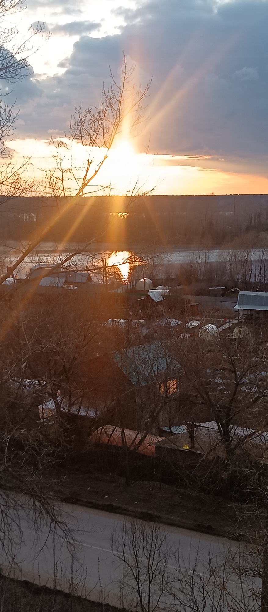The image presents a serene landscape captured during either sunrise or sunset, characterized by warm, golden light. The sun, a brilliant and intense source of light, is positioned prominently on the left side of the upper frame, emitting powerful crepuscular rays that fan out across the sky and through the scene. The sky is partly cloudy, with varying shades of grey and soft orange hues.
In the middle ground, a wide river or body of water reflects the sun's brilliance, creating a shimmering path of light across its surface. Beyond the river, a distant, dark treeline or forest stretches across the horizon.
Numerous bare-branched deciduous trees, indicative of late autumn, winter, or early spring, occupy the foreground and mid-ground, their intricate silhouettes adding depth and texture to the composition. Nestled amongst these trees and along the riverbank are several modest houses or small buildings, some with visible roofs. A warm, orange light glows from within the windows of some of these structures, suggesting internal illumination.
A narrow paved road with a faint white dividing line is visible in the bottom-left foreground, partially obscured by the foreground trees, whose reflections can be faintly seen on the wet-looking surface.
No people or specific activities are depicted in the image. The overall impression is one of quiet natural beauty, emphasizing the dramatic interaction of light and shadow during a transitional period of the day. There is no visible text.
