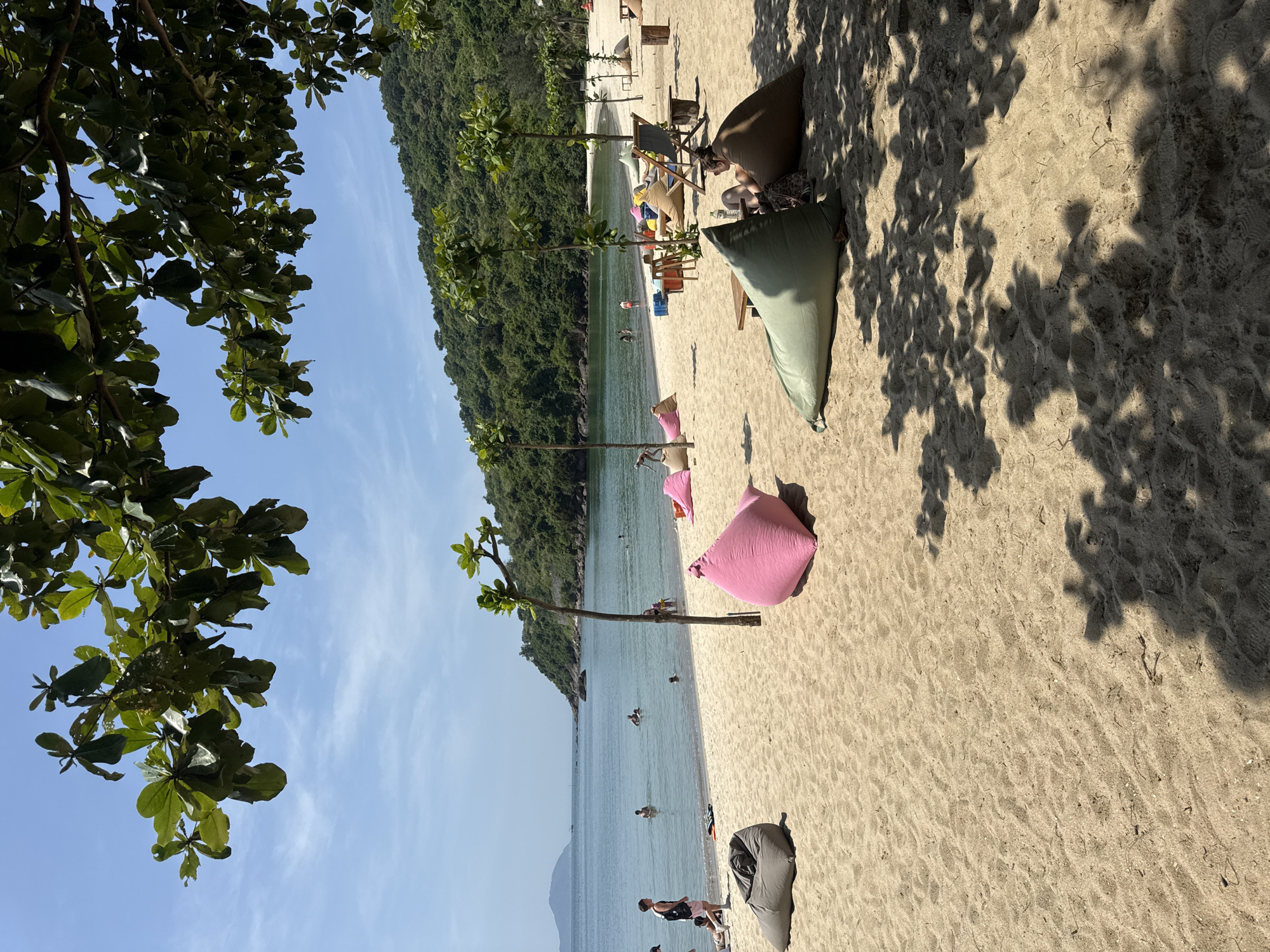 The image depicts a beach scene in an unknown location. The environment includes a sandy beach with scattered beanbag chairs of various colors, trees, and water. The water appears calm, with people swimming and standing in it. Lush green vegetation covers the land adjacent to the beach. Some people are on the beach, and shadows from trees are cast on the sand. The sky is clear blue.