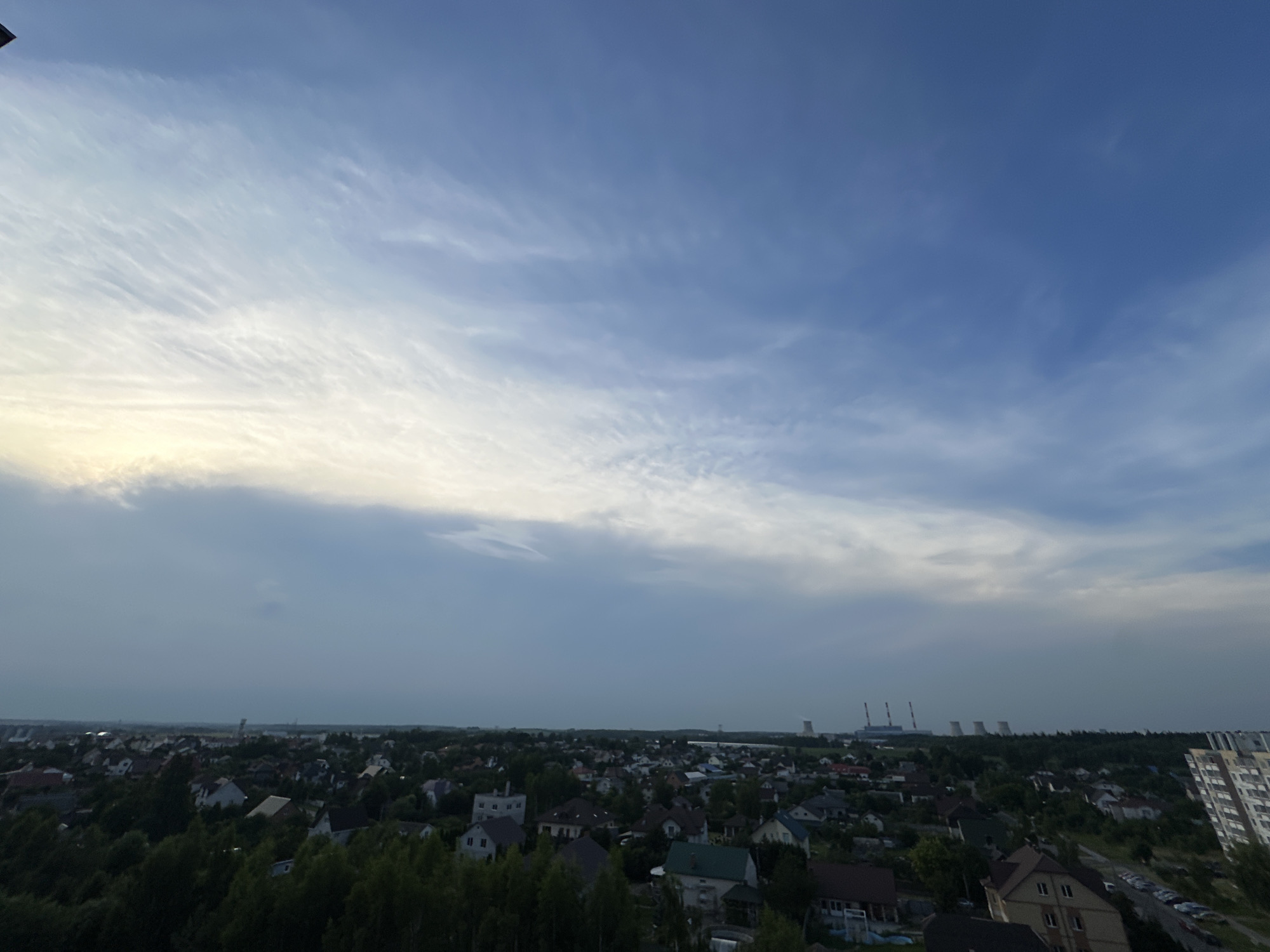 This is an aerial view of a suburban area in Шчомысліцкі сельскі Савет, Belarus, likely taken during the late afternoon or early evening, given the soft lighting and the sky's appearance. The scene is dominated by a vast expanse of sky, featuring a gradient of blue with wispy, white clouds streaking across the upper portion, transitioning to a more overcast and muted gray near the horizon.
Below the sky lies a sprawling residential area characterized by numerous houses with varied rooflines and colors, interspersed with dense green trees and vegetation. The buildings appear to be mostly detached homes, suggesting a suburban or semi-rural setting. In the distance, several industrial structures, including tall chimneys, are visible, indicating the presence of a power plant or similar industrial facility. A modern high-rise apartment building stands out on the right side of the frame, contrasting with the more traditional housing. The overall impression is one of a quiet, residential landscape under a dynamic sky. No people or specific activities are discernable in the image. No visible text is present.