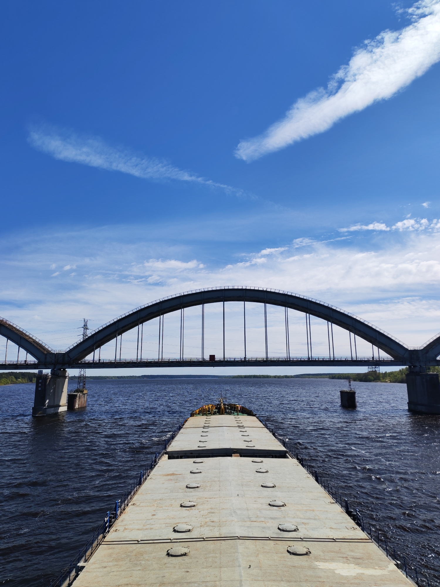 A wide-angle shot depicts a gray, deck barge traveling on the Volga River towards the arched bridge in Gladyshevo, Russia. The bridge's gray arches are supported by concrete piers set in the river. Two towers sit on either side of the central portion of the bridge, presumably for maintenance. The barge is flat and covered with a gray, concrete-like material, with circular access points. The dark water of the Volga stretches to the horizon, with visible waves and currents. The sky is a bright blue with wispy white clouds. Trees line the horizon on either side of the river.