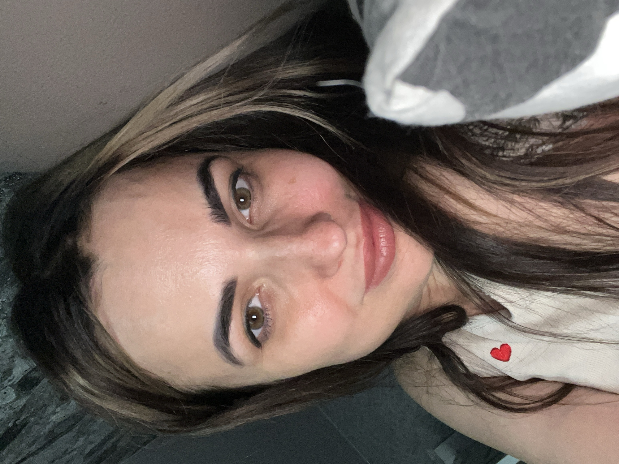 The image captures a close-up, tilted portrait of a woman. She has long, dark brown hair with lighter highlights, brown eyes, and defined eyebrows. Her lips are slightly parted in a soft smile, and she looks directly at the camera. Her skin appears clear with natural texture.
A grey and white striped or patterned fabric, possibly a blanket or clothing, partially covers the upper right side of the frame. On the lower right, a light-colored garment, potentially a top, is visible, featuring a small, embroidered red heart symbol. The background is largely out of focus and indistinct, with a dark, textured surface visible on the lower left.
The setting appears to be indoors, indicated by the soft, even lighting and lack of outdoor elements. The woman's direct gaze and expression suggest she is posing for a selfie or a close-up photograph. Her demeanor appears calm and pleasant. There are no explicit location cues, time of day indicators, or weather details visible. The notable detail is the small red heart embroidery on her clothing.