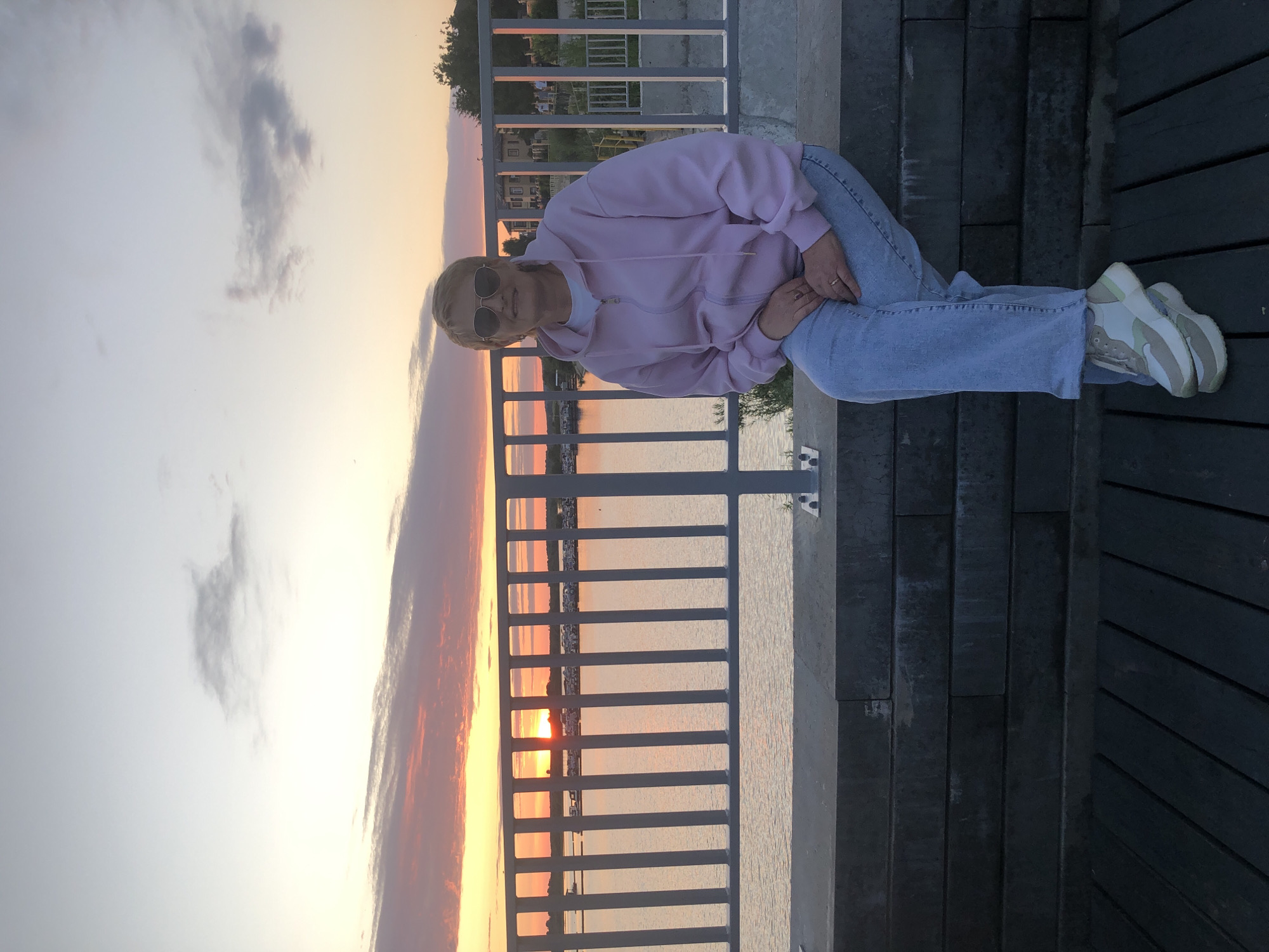 This vertical, full-body shot captures a person sitting on a wooden deck, leaning against a railing. The subject, with short blonde hair and wearing sunglasses, is dressed in a light pink hoodie, blue jeans, and white and light green sneakers. They are looking towards the camera with a relaxed expression.
Behind the railing, a body of water is visible, reflecting the warm hues of a sunset. The sky is filled with soft, diffused clouds, suggesting either early morning or late evening. The scene is set outdoors, possibly a waterfront promenade or park area, given the presence of the railing and wooden decking. The lighting indicates it is either sunrise or sunset, casting a soft glow over the landscape and the subject. The overall mood is tranquil and serene.
The location context of Solikamsk, Russia, is not directly evident from visual cues in the image itself. No visible text is present.