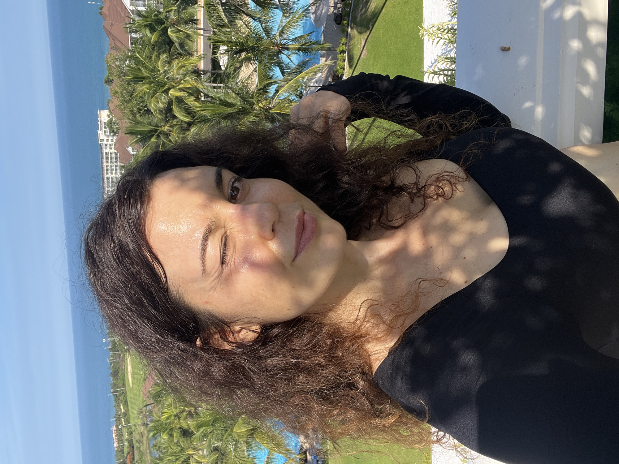 A woman with long, dark, curly hair is taking a selfie outdoors, winking at the camera. She is wearing a black, long-sleeved top. She is standing on a balcony or railing, with a view of palm trees, a swimming pool, buildings, and the ocean in the background. The sun is shining brightly, casting shadows on her face and clothing. The location is Phu Thuy Ward, Vietnam.