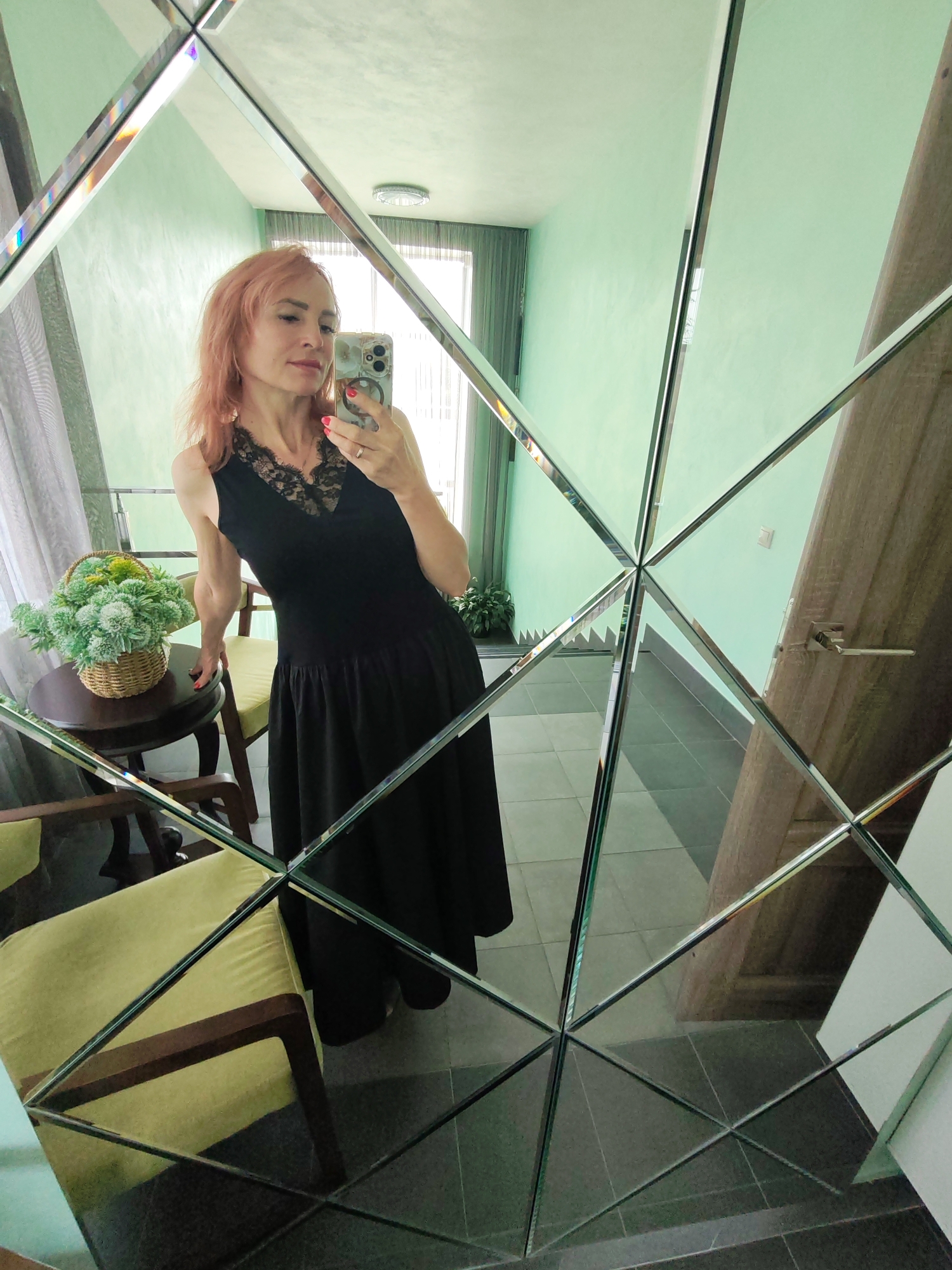 A woman with pink hair is taking a selfie in a mirrored wall. She is wearing a black dress with lace detailing and is holding a smartphone. She is standing in a hallway with mint green walls and a tiled floor. There is a table with a basket of artificial flowers to her left, and chairs in the foreground. The mirror is composed of multiple square panes, creating a faceted effect.