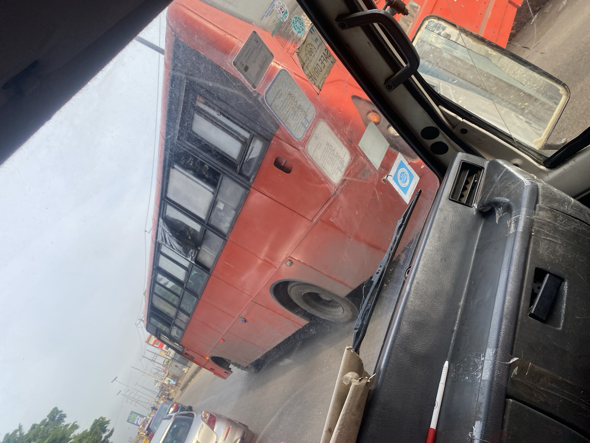 The image shows a view from inside a vehicle, looking out at a red bus stuck in traffic on a cloudy day in Ikorodu, Nigeria. The bus is large and appears to be a public transport vehicle. It has multiple windows and is the dominant subject in the frame. Other vehicles, including a silver car, are visible in the background, suggesting a congested road. The environment appears to be urban, with streetlights and buildings lining the road. The road surface is wet, indicating it may have rained recently or the area is prone to water accumulation. The dashboard of the vehicle from which the photo is taken is visible in the foreground, showing some wear and tear. There are some papers or documents attached to the side of the red bus, possibly indicating permits or registrations. The overall scene depicts a common urban traffic situation in Ikorodu.