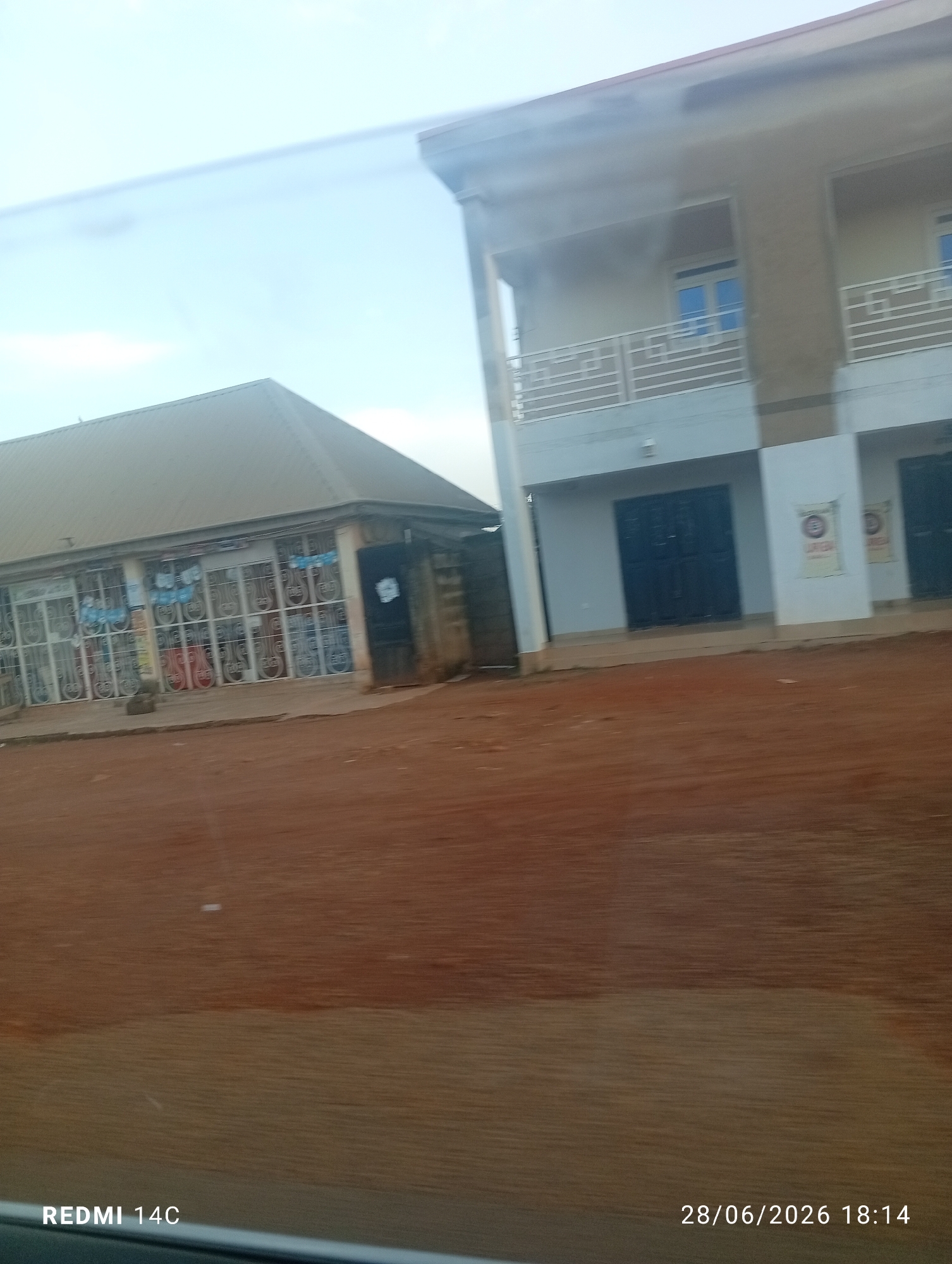 In the city of Jos, Nigeria, a street scene is depicted with two buildings visible. On the left, a single-story building with a pitched roof features a storefront with numerous windows displaying what appear to be musical instruments, possibly guitars, arranged in a grid. The windows are secured with metal grates. To the right, a multi-story building with a flat roof and balconies is under construction or recently completed. The lower level has dark doors, and the upper level has windows with blue glass and white railings on the balconies. The ground in front of the buildings is a reddish-brown dirt. The sky is overcast. A timestamp in the bottom right corner indicates the image was taken on 28/06/2026 at 18:14. The bottom left corner displays "REDMI 14C".