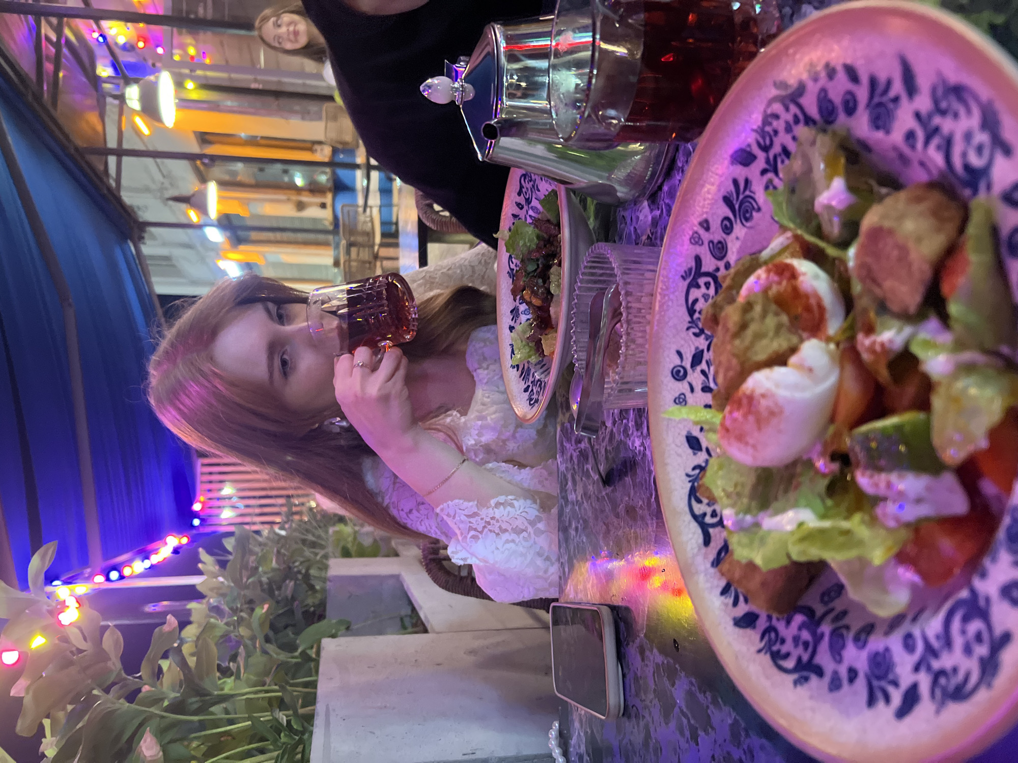 In this media file, a young woman with long, reddish-brown hair is seated at a table, drinking from a glass of dark liquid, possibly tea or a beverage. She is wearing a white lace top. The table is covered with a marble-like patterned surface and is adorned with two plates of food, one containing a salad with what appears to be pieces of meat, lettuce, and a creamy dressing, and another plate with a darker dish. A silver teapot and a glass teacup are also visible on the table.
The setting appears to be an outdoor or semi-outdoor dining area, possibly a restaurant or cafe, in the evening. String lights with red, blue, yellow, and white bulbs are visible in the background, creating a festive atmosphere. The overall lighting is dim, with colorful reflections on the table, suggesting the presence of decorative lights. In the background, another person with a smiling face is partially visible. The foliage in the foreground adds to the outdoor ambiance. The style of the plates, with their blue and white patterned rims, might suggest a particular cuisine or decor theme. The presence of a mobile phone on the table suggests it might be a casual outing. The image was taken in Nizhny Novgorod, Russia.