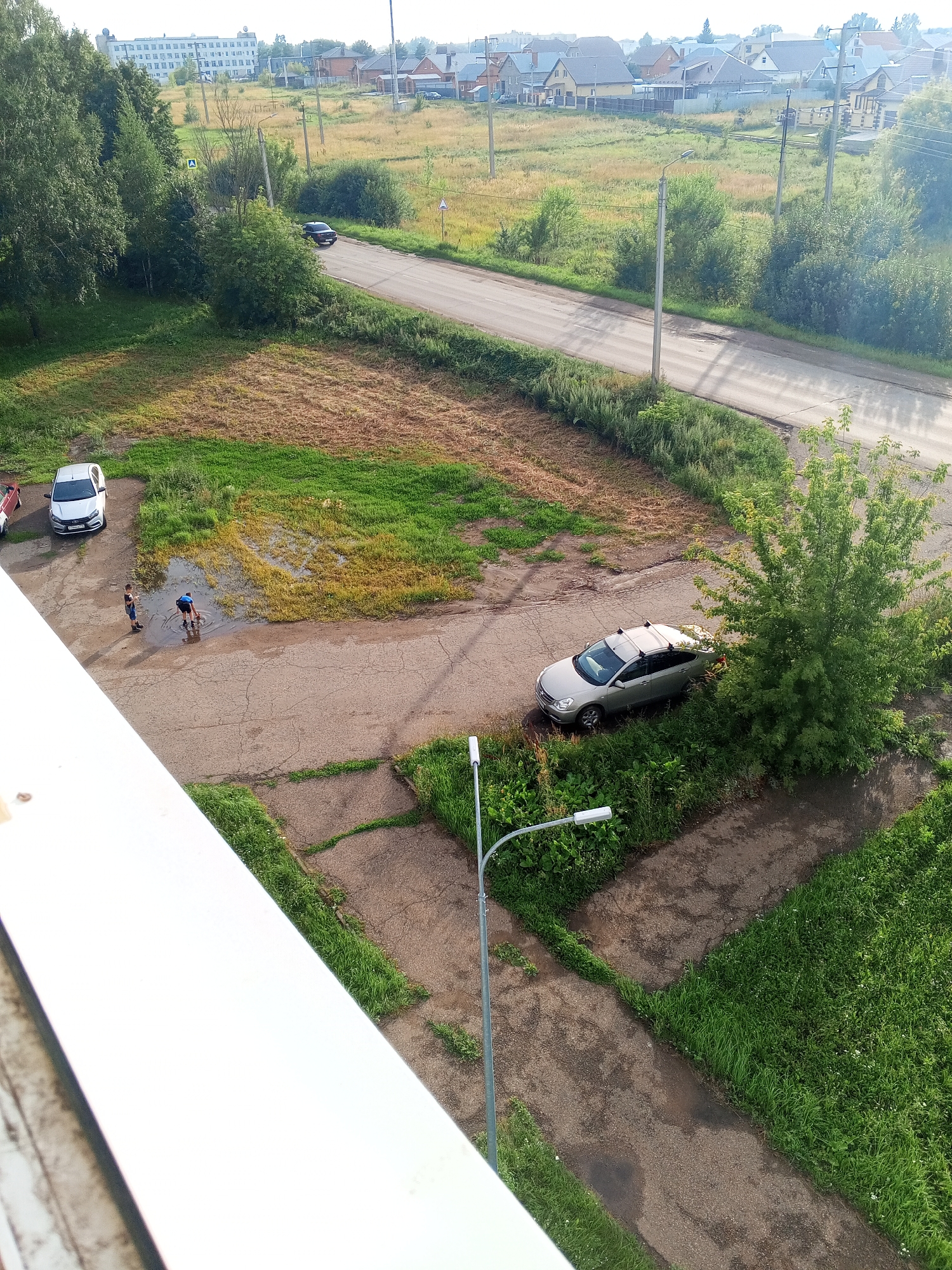 The image shows a residential area in Chistopol, Russia, with a road, several cars, two individuals, and various trees and vegetation. In the foreground, a white balcony railing is visible. On the road, a silver sedan is parked next to a cluster of trees. Further down the road, a white car is parked, and two individuals are standing near a large puddle of water, with one person appearing to interact with the water. The background reveals a street with other houses and a large building in the distance, surrounded by green fields and trees. A street light is positioned on the right side of the frame.