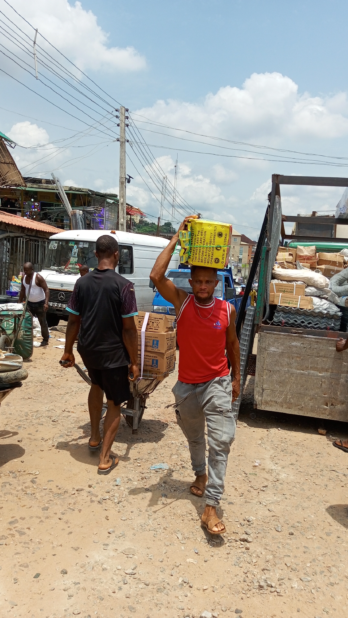 This outdoor photograph captures a bustling scene on a dirt road, likely a market or commercial area in Ogbunike, Nigeria, under a bright, cloudy sky, suggesting daytime. The main subject is a man in the foreground, wearing a red tank top, grey cargo pants, and brown sandals, carrying a yellow cardboard box on his head. He has a focused, neutral expression and looks directly at the camera. The box on his head displays text, including "ERSTAR E6013", a warning about equipment use, and "G.W/N.W: 21/20KG", indicating a net weight of 20kg.
Behind him, another man, seen from the back, wears a black t-shirt and shorts, pushing a metal wheelbarrow loaded with several brown boxes. In the background, a white Mercedes-Benz van is parked, and a blue vehicle is partially visible. To the far right, part of a truck bed, heavily loaded with various goods like sacks, boxes, and what appears to be metal mesh or grating, is visible. Overhead, numerous power lines stretch across utility poles, indicating an urban or semi-urban environment. The overall scene depicts men engaged in the manual transportation and distribution of goods.