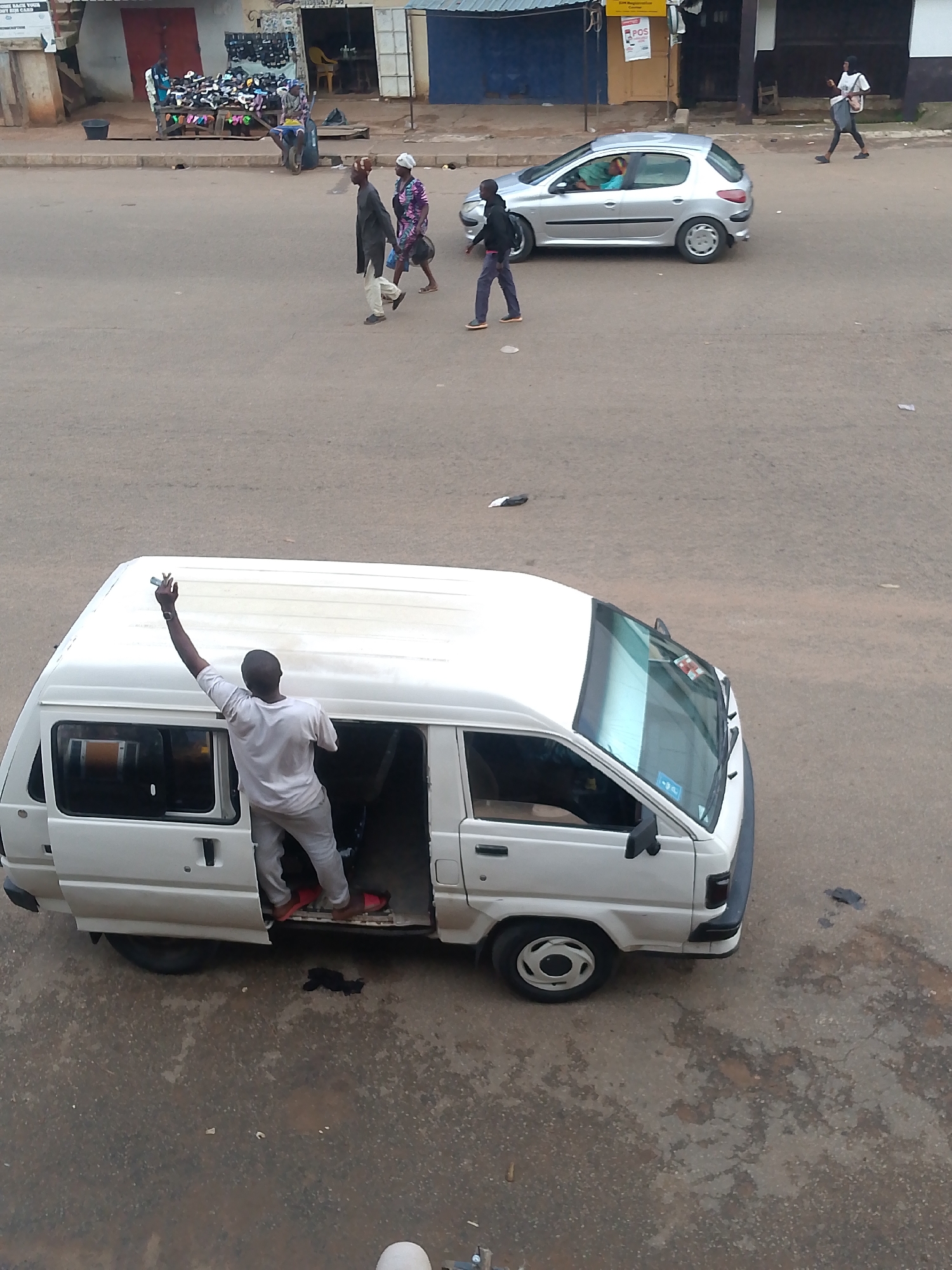 In Bukuru, Nigeria, a man wearing a white t-shirt and light-colored pants stands with his back to the camera, reaching up with his right arm and holding a white object. He appears to be interacting with the roof of a white van. The van has its driver's side door and a rear sliding door open. Inside the van, a person wearing red sandals can be seen. The van is parked on a paved road, and in the background, a silver car drives past. Further in the distance, a shoe stall is visible, with several people walking around it. The scene depicts everyday street activity in a Nigerian city.