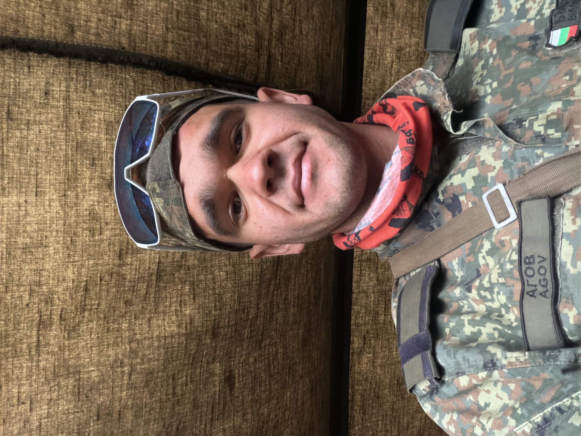 A man wearing a camouflage uniform, a camouflage cap, and sunglasses is taking a selfie. He has a red bandana around his neck. A nametag on his uniform reads "АГОВ / AGOV". The background is a textured brown surface, likely canvas or fabric. The image appears to be taken outdoors. There is a Bulgarian flag patch on the man's left shoulder.