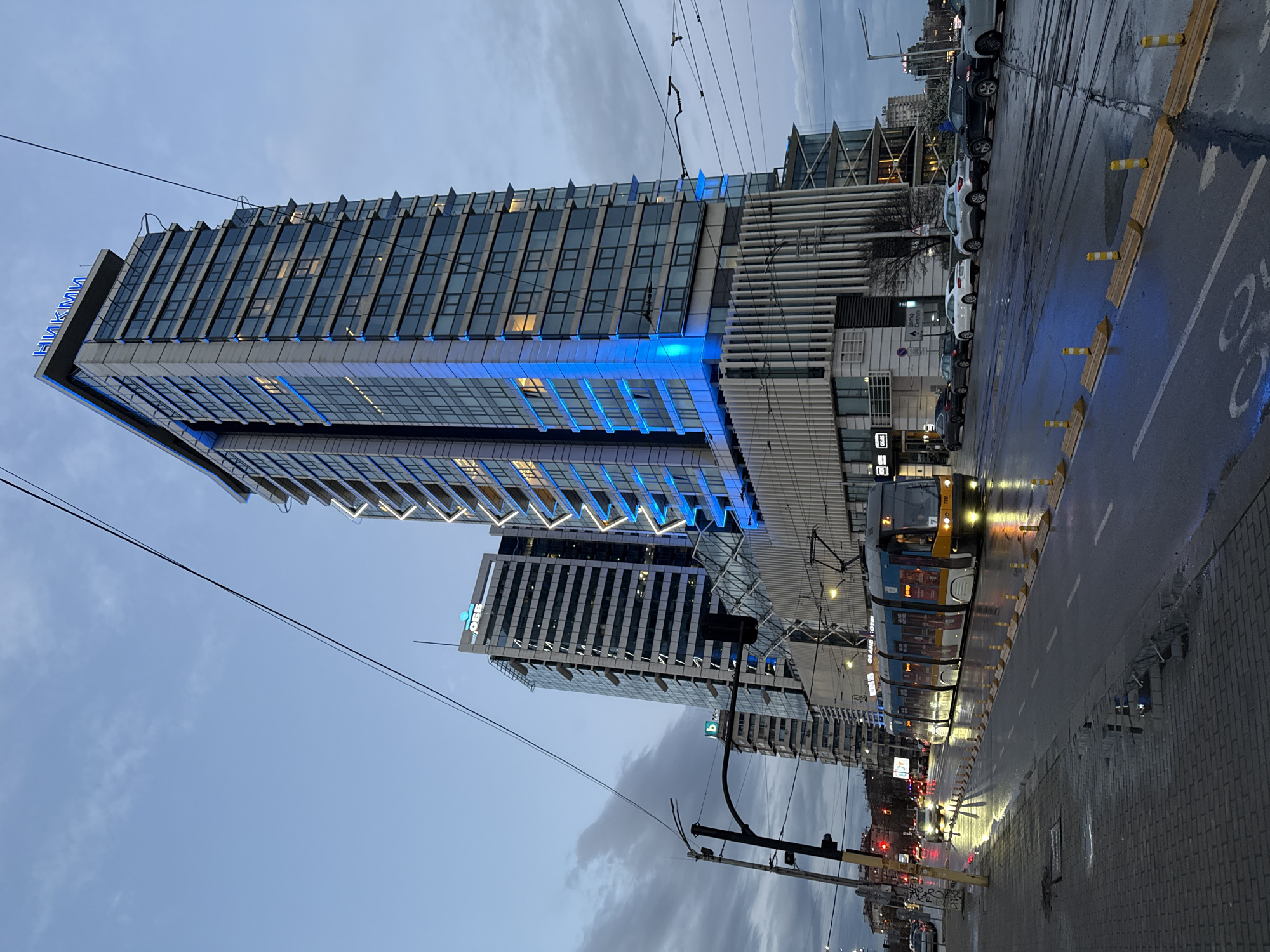 The image shows a cityscape in Sofia, Bulgaria, at dusk or dawn on a wet day. The focus is on a tall, modern building with blue lighting accents, which appears to be a hotel, possibly part of the Hyatt Regency Sofia complex, identified by the Cyrillic lettering "НИКМИ" at the top. Another skyscraper, labelled "ОББ", is visible in the background. A tram is present in the scene, traveling on tram tracks along the wet road. The street is lined with parked cars and yellow safety bollards. The sky is overcast. Overhead electrical lines for the tram cross the image. The perspective is from street level, tilted slightly upward toward the buildings. The road and sidewalk appear wet, reflecting the ambient light and traffic signals.
