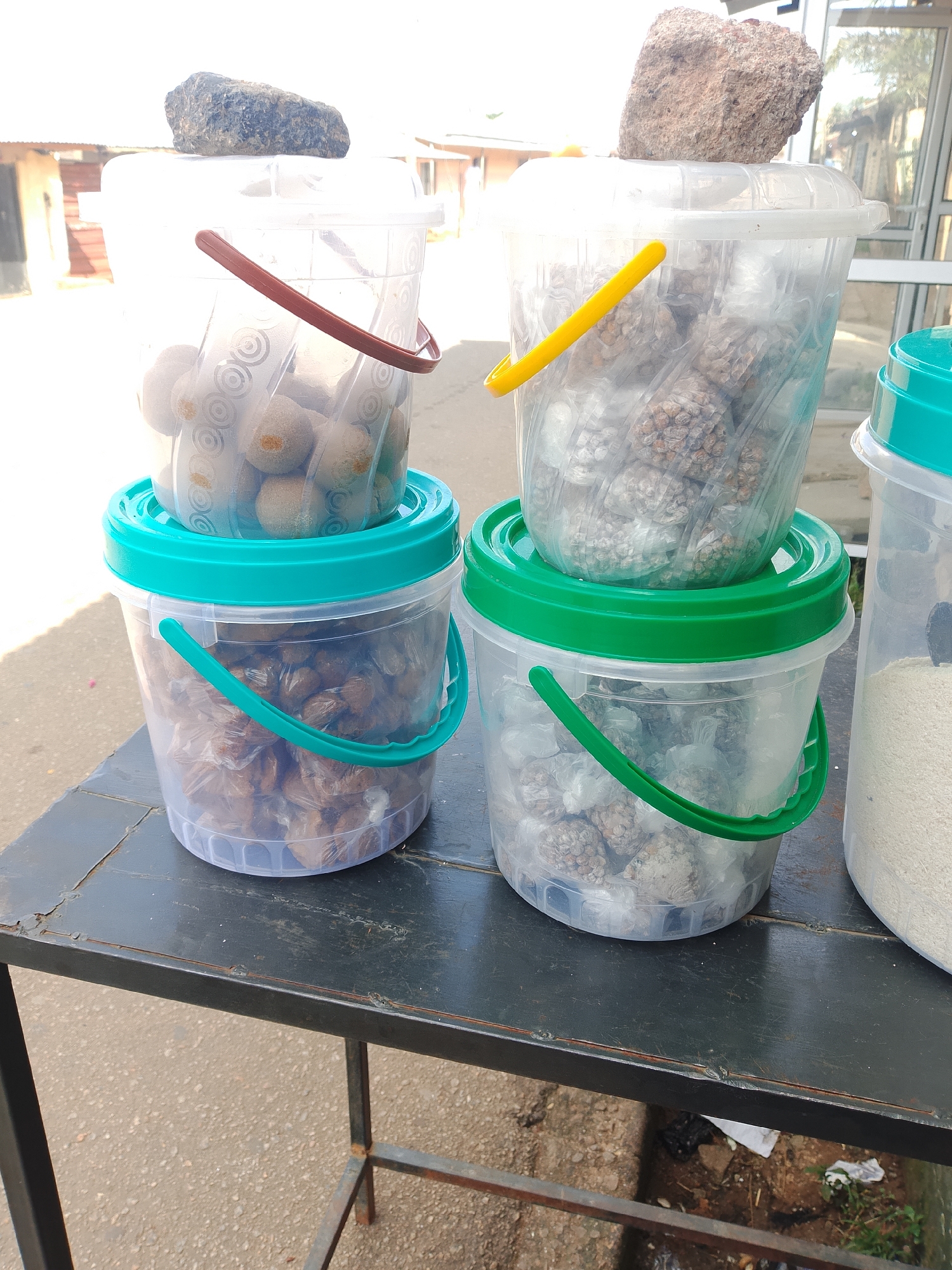 Four clear plastic buckets with green or brown handles and lids are stacked and arranged on a dark metal table. The two buckets in the foreground are stacked, with the bottom bucket filled with small, individually wrapped brown items, and the top bucket on that one is filled with round, light brown items in clear plastic wraps. To the right of these stacked buckets, another clear plastic bucket with a green lid contains small, individually wrapped light brown clusters. To the far right, a fourth clear plastic bucket with a green lid is filled with a fine white granular substance. Each of the top three buckets has a rock placed on its lid. The table is situated outdoors on a paved surface, with a building and vegetation visible in the background. The lighting suggests it is daytime.