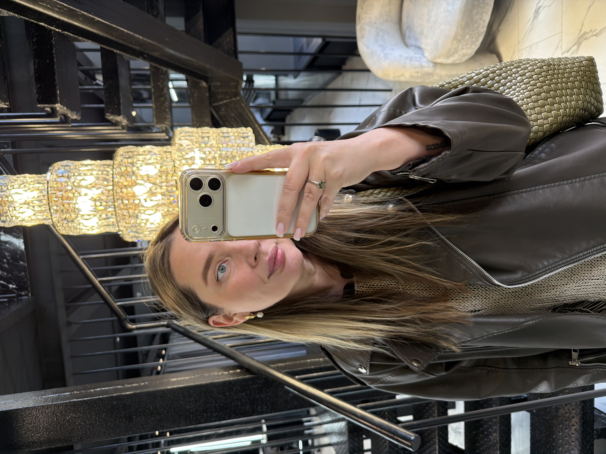 An indoor, selfie-style portrait of a woman, taken in a setting suggestive of a modern building or upscale interior, possibly in Moscow, Russia. The image is currently rotated 90 degrees clockwise. The subject is a woman with light brown and blonde hair, blue eyes, and fair skin, looking directly at the camera with a calm, composed expression. She is wearing a dark olive or brown leather-like jacket over a lighter, possibly knitted, top.
She holds an iPhone in a clear case with her right hand, positioned as if taking a selfie. On her left ring finger, she wears a ring with a prominent green stone. A pearl ear cuff is visible on her right ear, and a tattoo, which appears to read "COCO," is visible on her inner left wrist. A woven olive-green bag rests on her right shoulder.
The background features a large, ornate chandelier composed of multiple tiers of rectangular, crystal-like elements, emitting a warm, bright light. Behind her, a dark metal staircase with visible railings and steps extends upwards, indicating a multi-level interior space. A light-colored, abstract sculpture or decorative element is partially visible in the upper right background. The scene suggests a stylish and well-lit environment.