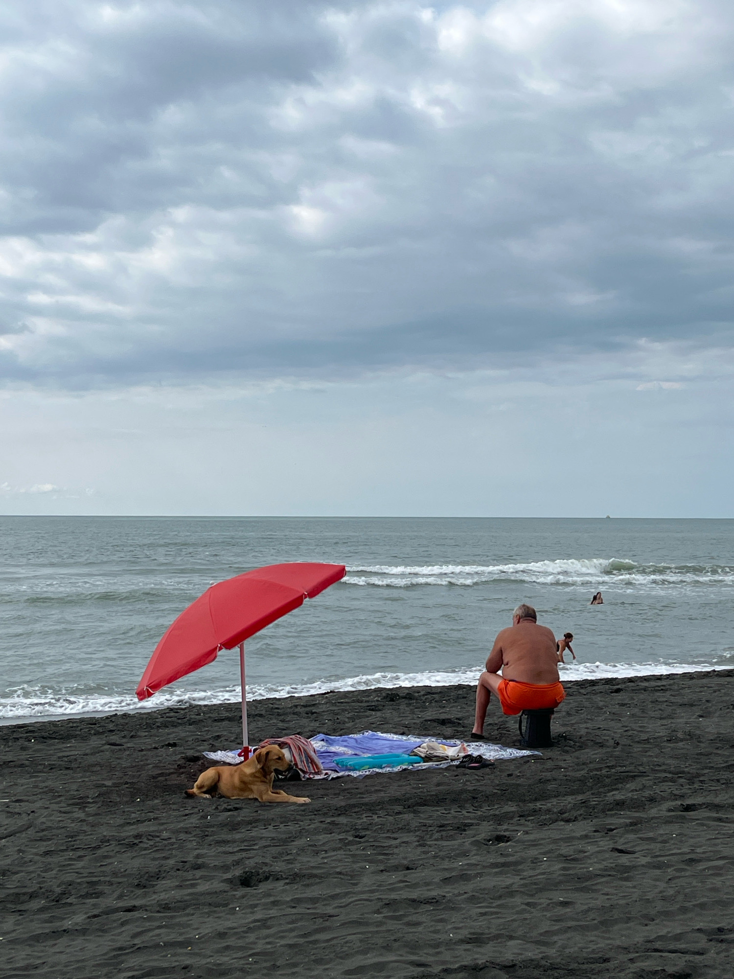 The image depicts a cloudy day at Ureki Beach in Georgia. In the foreground, a brown dog lies on the black sand near a red umbrella. A person in orange shorts is seated on a dark object facing the sea. The sea is choppy with white-capped waves. In the background, two people are visible in the water, and a distant boat can be seen on the horizon. The sky is filled with heavy, gray clouds.
