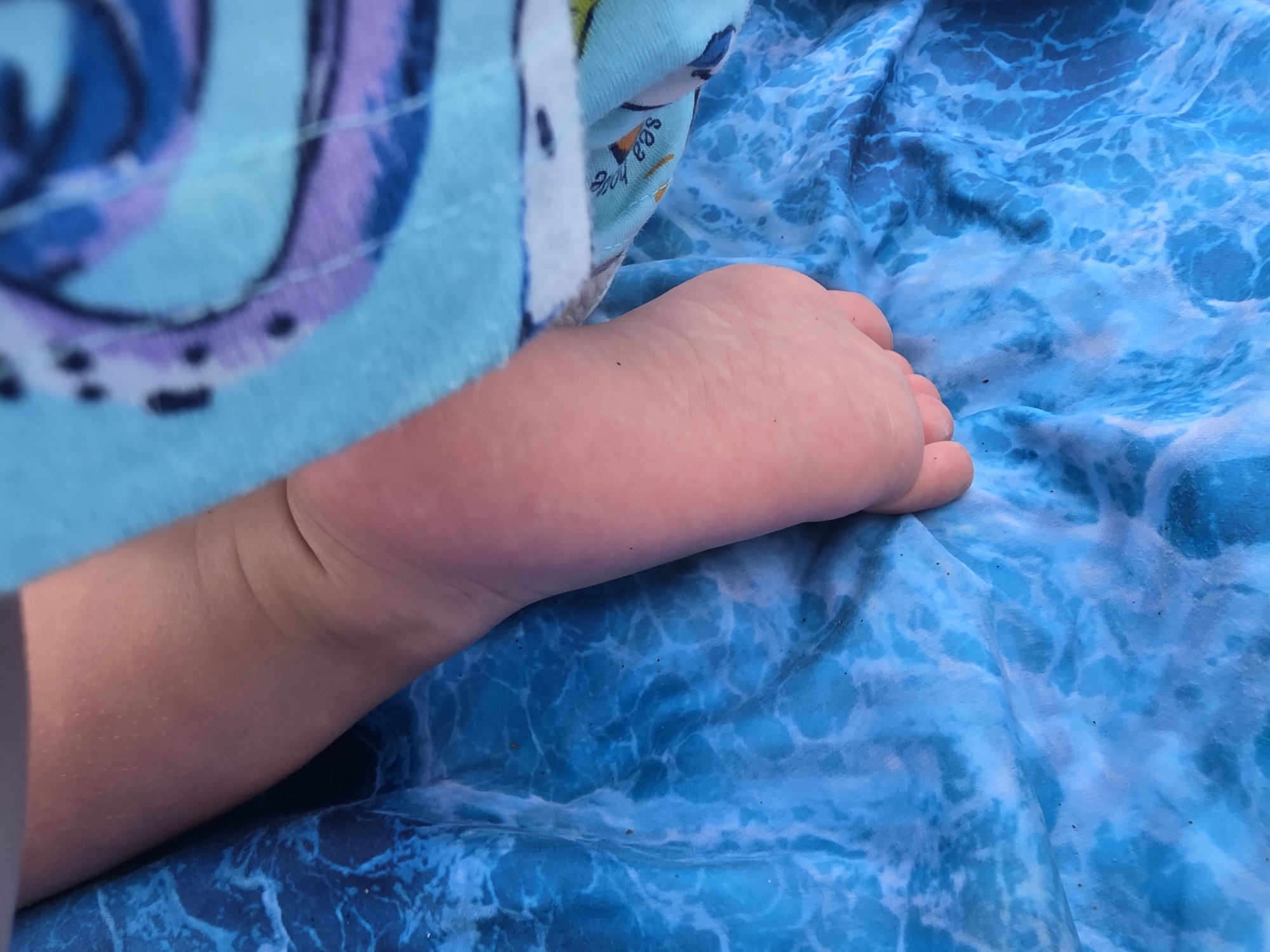 A close-up shot focuses on the bare foot and lower leg of a young child, resting on a blue fabric with a water-wave pattern. The child's skin is smooth and fair. A colorful towel or blanket, featuring a swirly pattern in shades of blue and purple, is draped over the child's leg. Some faint text is visible on the edge of this fabric, appearing to say "sea hog" or something similar. The background fabric is a bright blue with a dynamic, rippling wave design, suggesting an outdoor setting possibly near water, such as a beach or poolside. The lighting suggests it is daytime, with no specific weather conditions apparent. The overall impression is one of a relaxed, possibly vacationing, scene. The child's foot is positioned on the right side of the frame, with the leg extending towards the left. Small dark specks, possibly dirt or sand, are visible on the blue fabric.