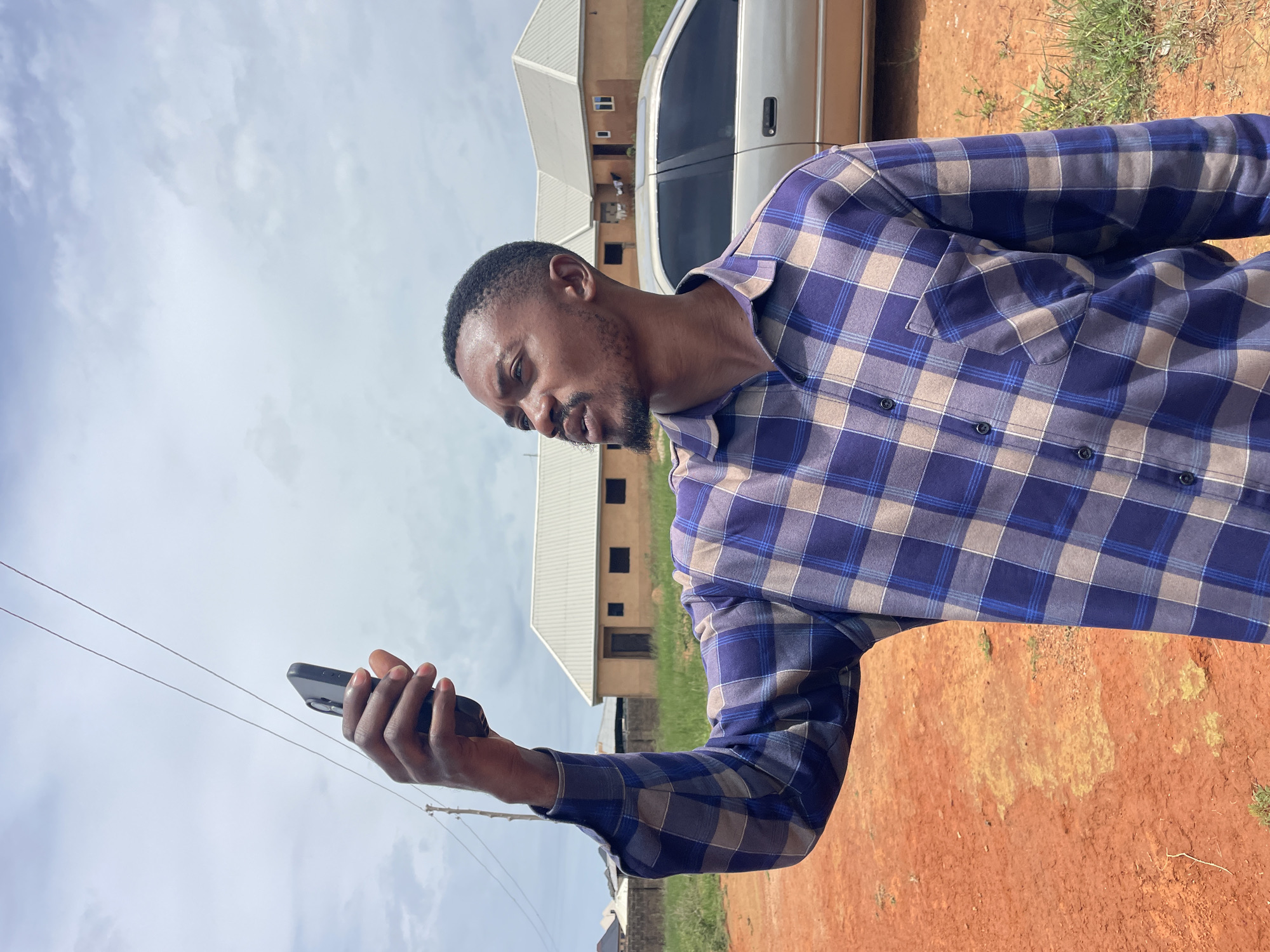 In the city of Rafin Bauna, Nigeria, a young Black man stands outdoors, holding a dark-colored smartphone aloft in his right hand, seemingly attempting to capture a photo or video. He is wearing a long-sleeved, plaid shirt with a pattern of blue, white, and grey squares. The man has a short haircut and a goatee.
The background reveals a slightly overcast sky with scattered clouds. Beneath the sky, two thin, parallel power lines stretch horizontally across the frame, supported by a utility pole on the left. Further in the background, several low-rise buildings with beige walls and white roofs are visible. A light-colored sedan is parked behind the man, positioned to his right.
The ground in the foreground is a reddish-brown, dusty earth, with patches of sparse green grass growing in places. The overall setting appears to be a somewhat undeveloped area on the outskirts of a town.