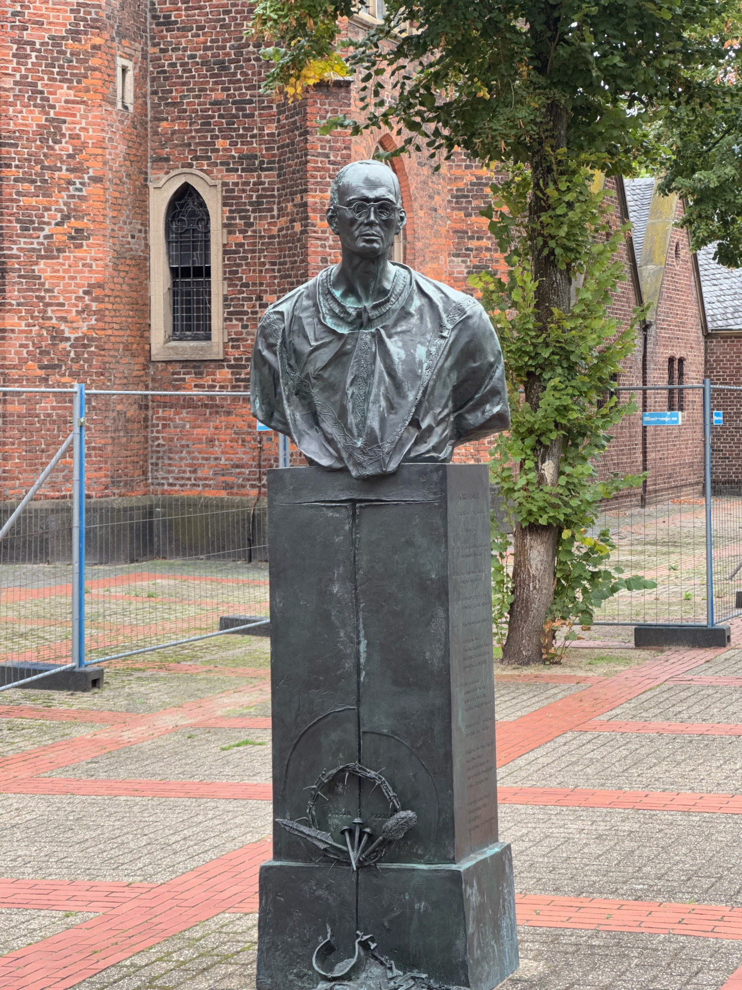 A full-body, eye-level, medium shot captures a bronze bust of a man, identified as Bishop Heinrich Janssen, on a tall, dark pedestal. The bust is positioned centrally in the frame, facing directly towards the viewer. The man in the bust wears glasses and has a bald head. He is dressed in clerical attire, likely a robe or cassock. The pedestal features engraved text and a relief of a crown of thorns with nails.
The bust and pedestal are situated outdoors on a paved area composed of alternating squares of light grey concrete and red bricks. In the background, a red brick building, possibly a church due to the Gothic-style window visible on its facade, is present. The building appears to be under construction or renovation, indicated by temporary metal fencing with blue plastic barriers surrounding the area. Trees with green foliage grow behind the bust and to the right of the building. The sky is overcast, suggesting a cloudy day.
The bust is located in front of the St. Lambertus Catholic Church in Cleves, Germany, specifically in the St. Lambertus churchyard. This location is confirmed by the presence of the church building and the historical context of Bishop Heinrich Janssen's association with the church.