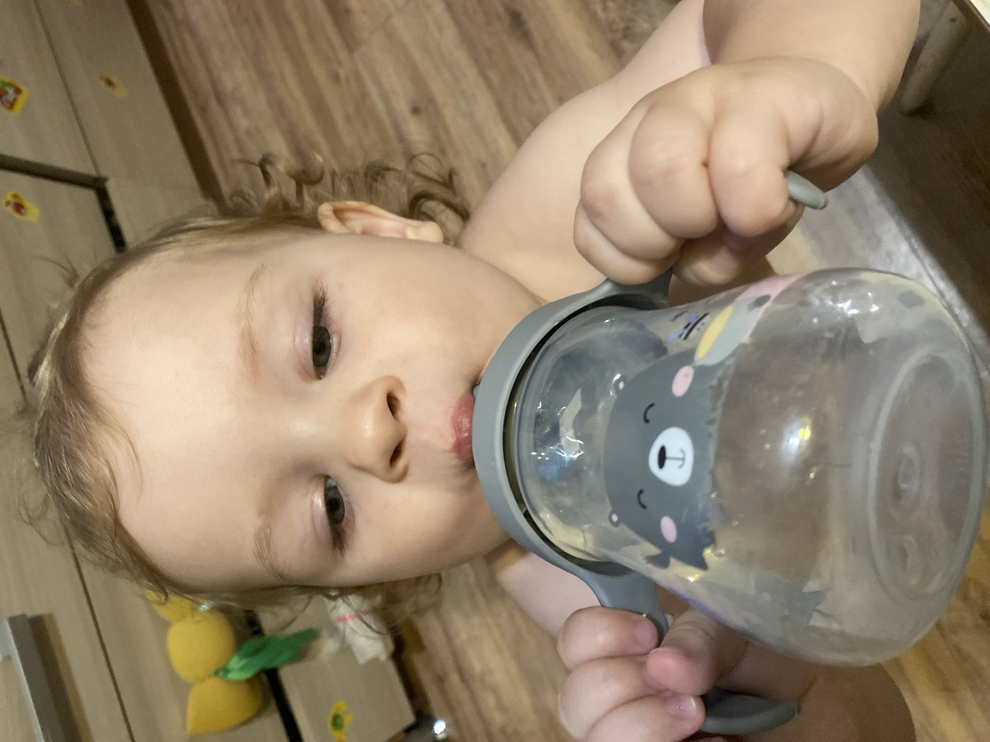 A close-up, high-angle, indoor shot shows a toddler drinking from a sippy cup. The toddler, who has light curly hair, is looking down at the cup. The cup is clear with a gray lid and handles, decorated with a smiling bear face. The background consists of a wooden floor and what appears to be a light-colored dresser or cabinet with yellow stickers. The lighting is dim, with some glare on the cup.