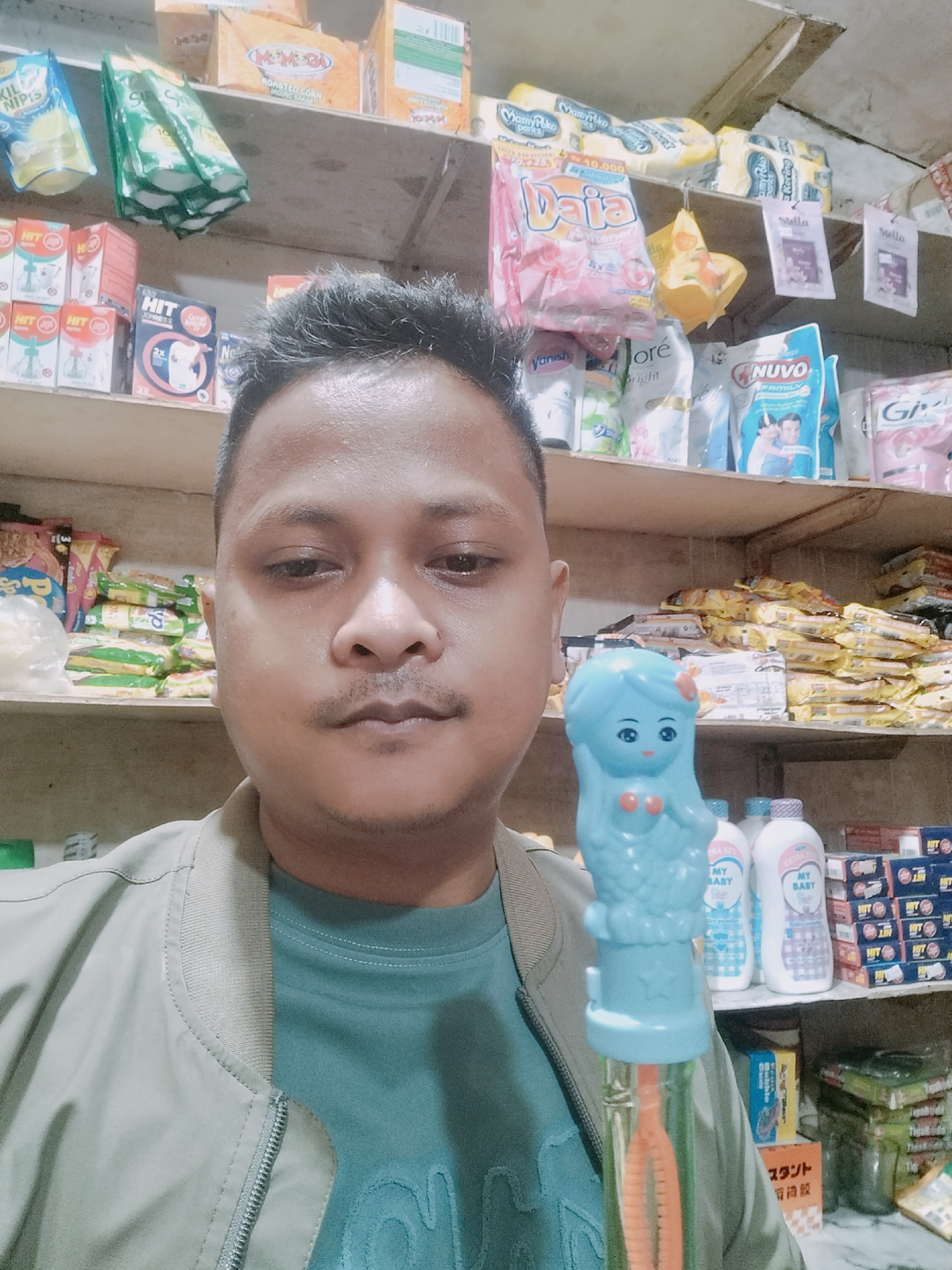 In Lhokseumawe, Indonesia, a young man with dark hair and a mustache poses for a selfie in what appears to be a small convenience store. He is wearing a light-colored bomber jacket over a teal t-shirt. In his right hand, he holds up a blue bubble toy shaped like a mermaid, with its transparent orange bubble wand visible. The background is filled with shelves stacked with various packaged goods, including detergents, snacks, and personal care items, suggesting a retail environment.