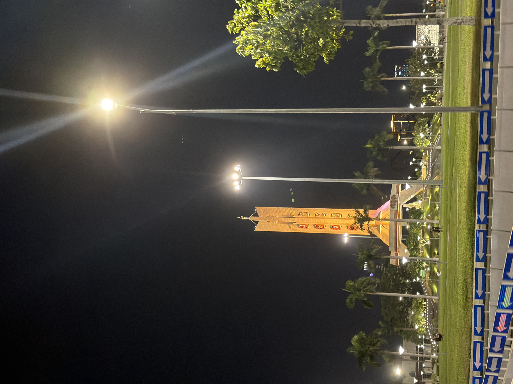This is a nighttime, low-angle, wide shot of the Dragon Bridge in Da Nang, Vietnam. The bridge, a prominent landmark, is illuminated and dominates the background. Several streetlights are visible, with one in the foreground emitting a strong glare that obscures a portion of the sky. The area around the bridge appears to be a public space with grass, palm trees, and other foliage. There are a few people scattered throughout the park-like setting. The image also features directional arrows on a blue and white barrier in the foreground, suggesting a roadway or pedestrian pathway. The overall scene captures the bridge illuminated at night, with the surrounding urban environment visible in the background.