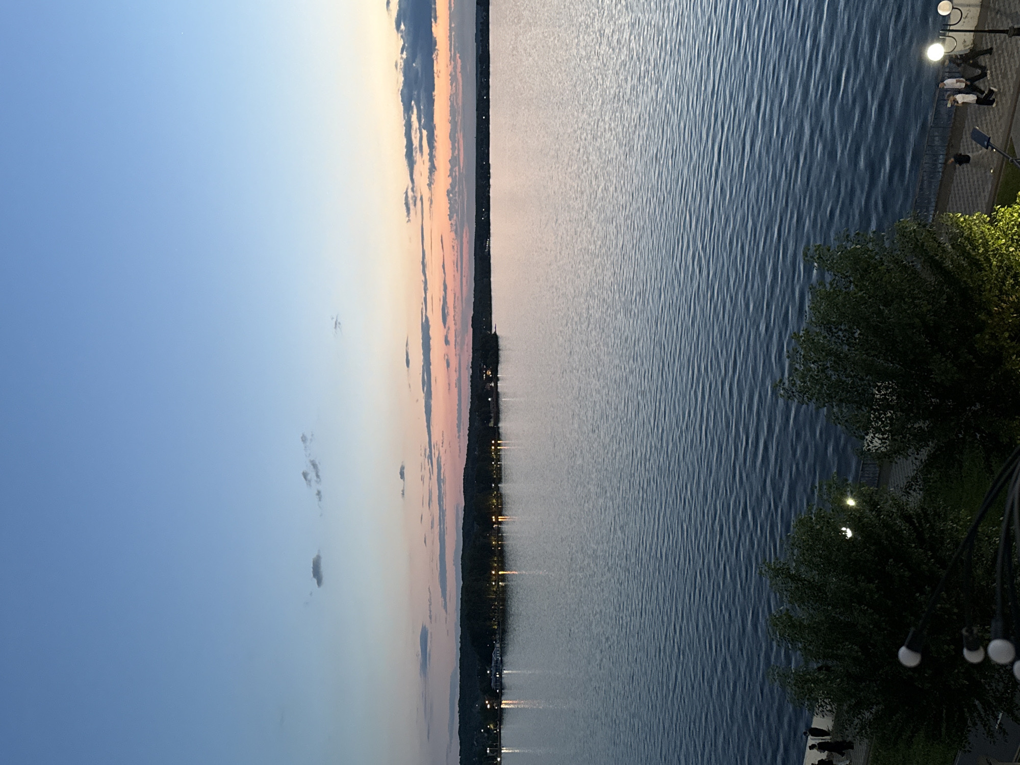 A serene wide-angle view captures a twilight scene over a large body of water in Votkinsk, Russia. The image is composed of three main horizontal sections: the sky, the water, and the waterfront promenade.
The sky, occupying the upper portion, displays the soft hues of dusk, transitioning from a clear, pale blue at the top to warmer shades of orange and pink closer to the horizon, with a few scattered, dark clouds.
The expansive body of water fills the mid-ground, its surface marked by gentle ripples that catch and reflect the fading light and the numerous artificial lights from the distant shore. This far shore, a dark silhouette against the evening sky, is dotted with dozens of bright, glowing lights from what appears to be a town or city. These lights create shimmering vertical reflections on the water, adding depth to the scene.
In the foreground, a paved promenade runs alongside the water. Several streetlights with luminous spherical bulbs cast warm light onto the patterned pavement and the dark green foliage of trees that line the walkway, some partially obscuring the lower right. Several people are visible on the promenade, standing or walking casually, enjoying the evening atmosphere. They appear as indistinct figures, suggesting leisurely activity. A dark railing separates the promenade from the water.
The overall mood is peaceful and tranquil, characteristic of a pleasant evening by the water. There is no visible text in the image.
