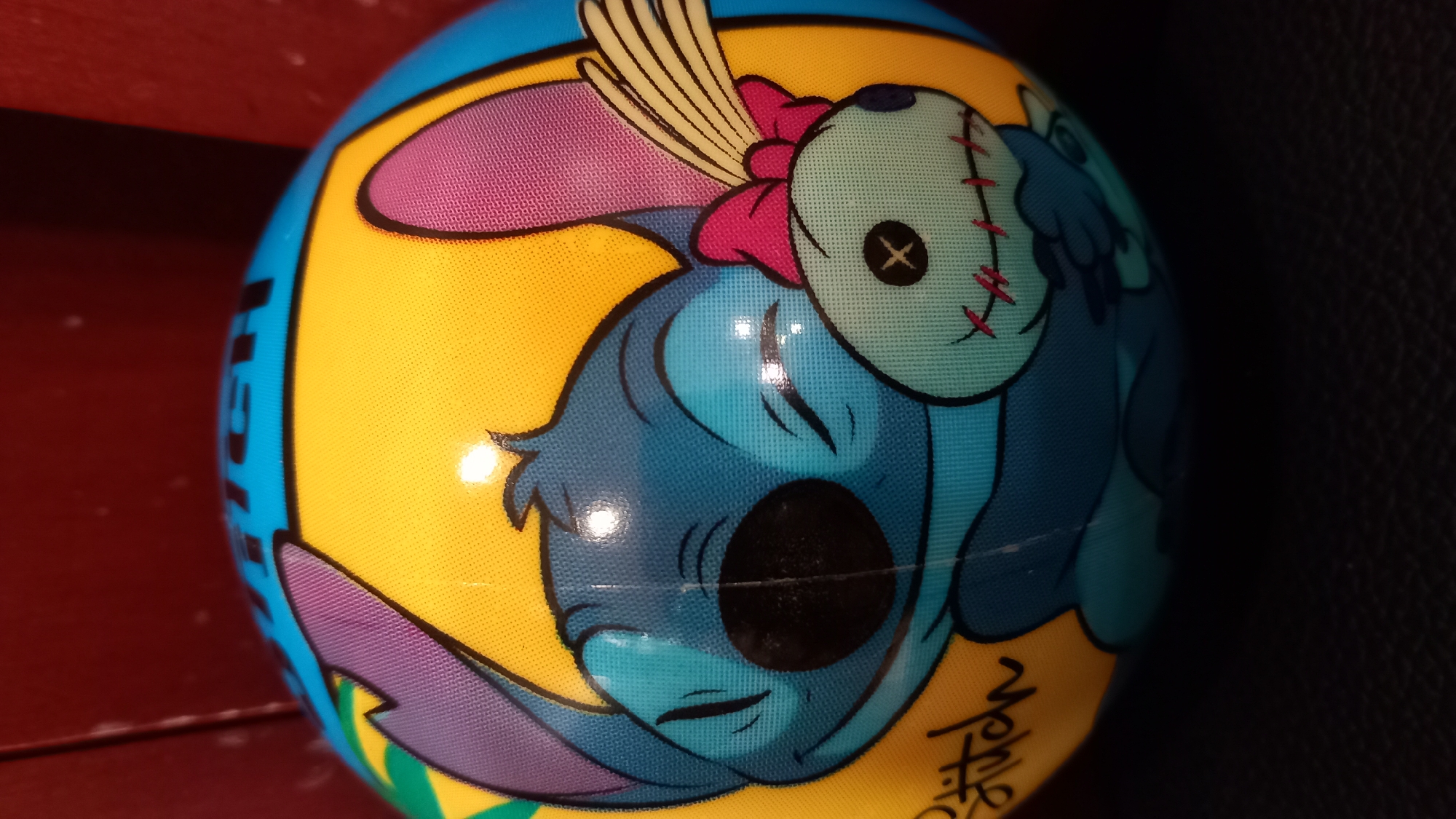 A close-up, slightly angled shot features a ball with a cartoon character, likely Stitch from Lilo & Stitch, prominently displayed. The character is blue with large, pointed ears, one purple and one pink. Stitch is depicted with closed eyes and a smile, hugging a light blue doll-like figure with stitch marks and a pink bow. The ball has a yellow background with blue and green accents, and the word "Stitch" is visible in black script on the lower left. The background is dark and blurred, with a hint of a reddish-brown surface above the ball. The scene is indoors and does not provide any information about a specific location in Kos, Greece.