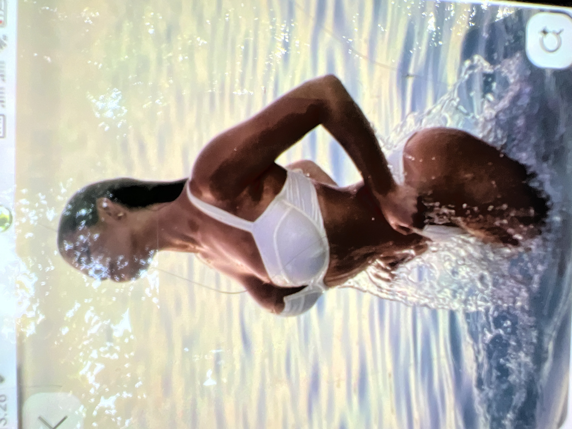 A woman in a white bikini stands in the water. Her head is turned to the right, and she is looking away from the camera. The water is rippling around her, and there are splashes of water on her skin. The background is a blur of light and color, suggesting a bright, sunny day. The image appears to be taken on a mobile phone, as evidenced by the UI elements at the top and bottom of the frame.