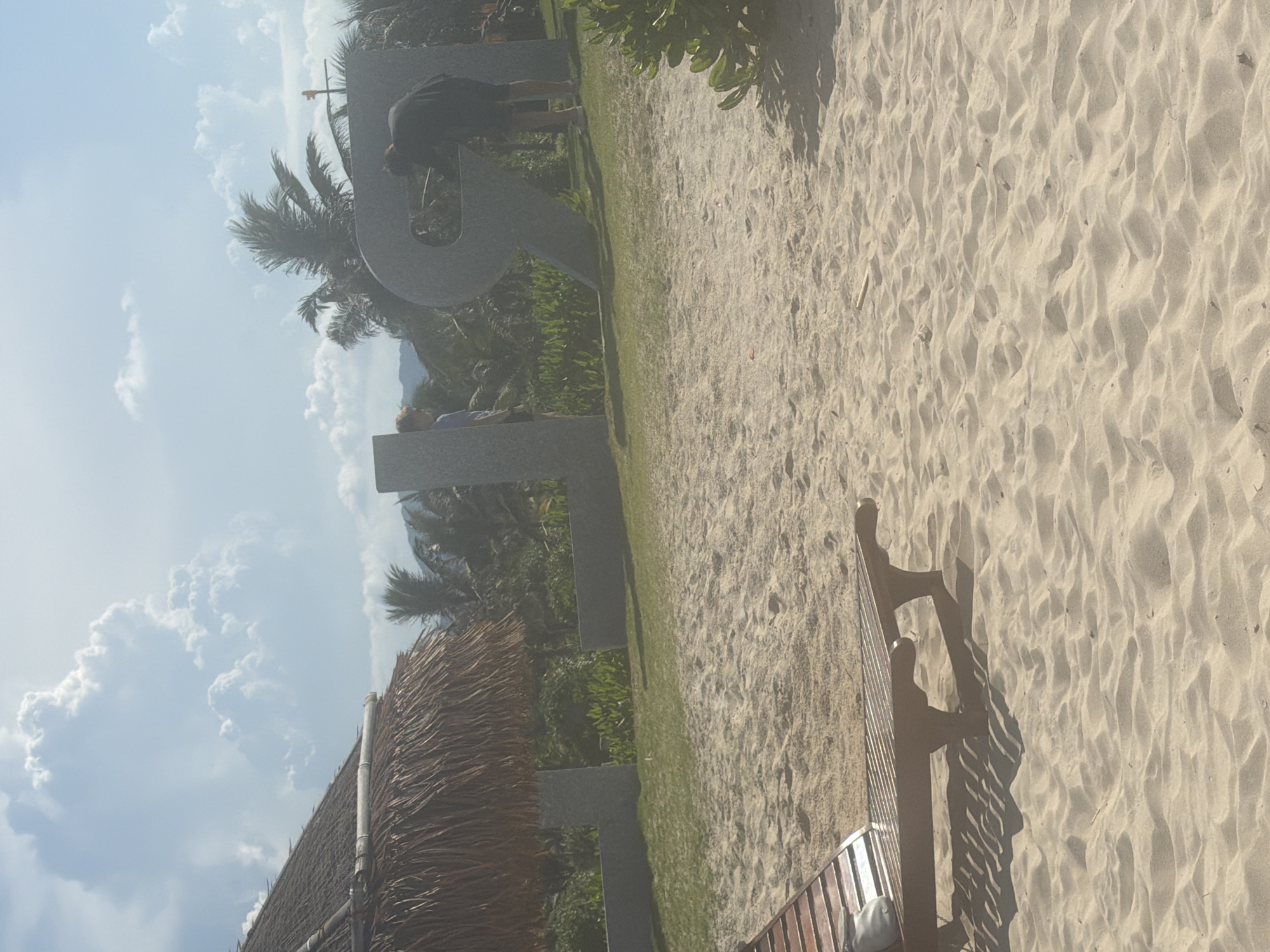 A sunny day in Bac Cam Ranh Ward, Vietnam, with a beach scene. In the foreground, a wooden lounge chair is positioned on the sand. Behind it, large concrete letters spelling out "LOVE" are visible on a grassy area. A person in a dark shirt and shorts is interacting with the letter "R," appearing to be cleaning or maintaining it. Another person, partially visible, stands near the letter "O." A thatched-roof structure, possibly part of a resort or building, is to the left, and lush green vegetation, including palm trees, forms the background. The sky is blue with scattered white clouds.