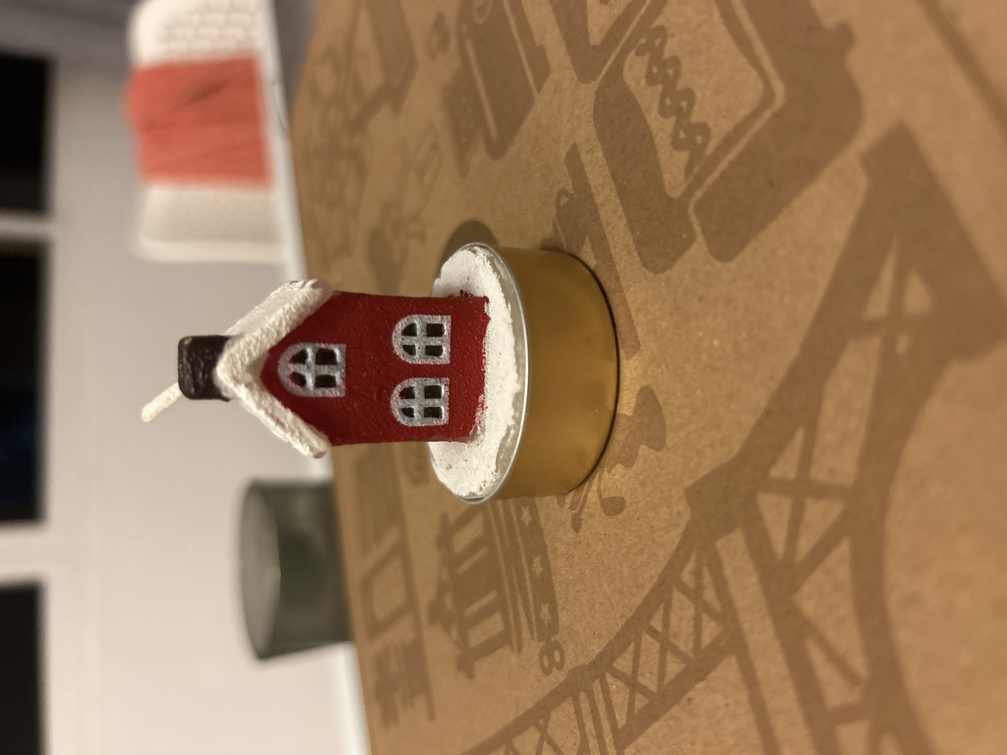 A close-up, angled shot focuses on a red, miniature house-shaped candle with white "snow" trim, embedded in a gold-colored tea light holder. The tea light is positioned in a hole within a light brown cardboard surface, decorated with outlined images including the Eiffel Tower. A window and the side of another cylindrical object are visible in the blurred background. The location, Câmara de Lobos, Portugal, cannot be determined from the visual information provided.