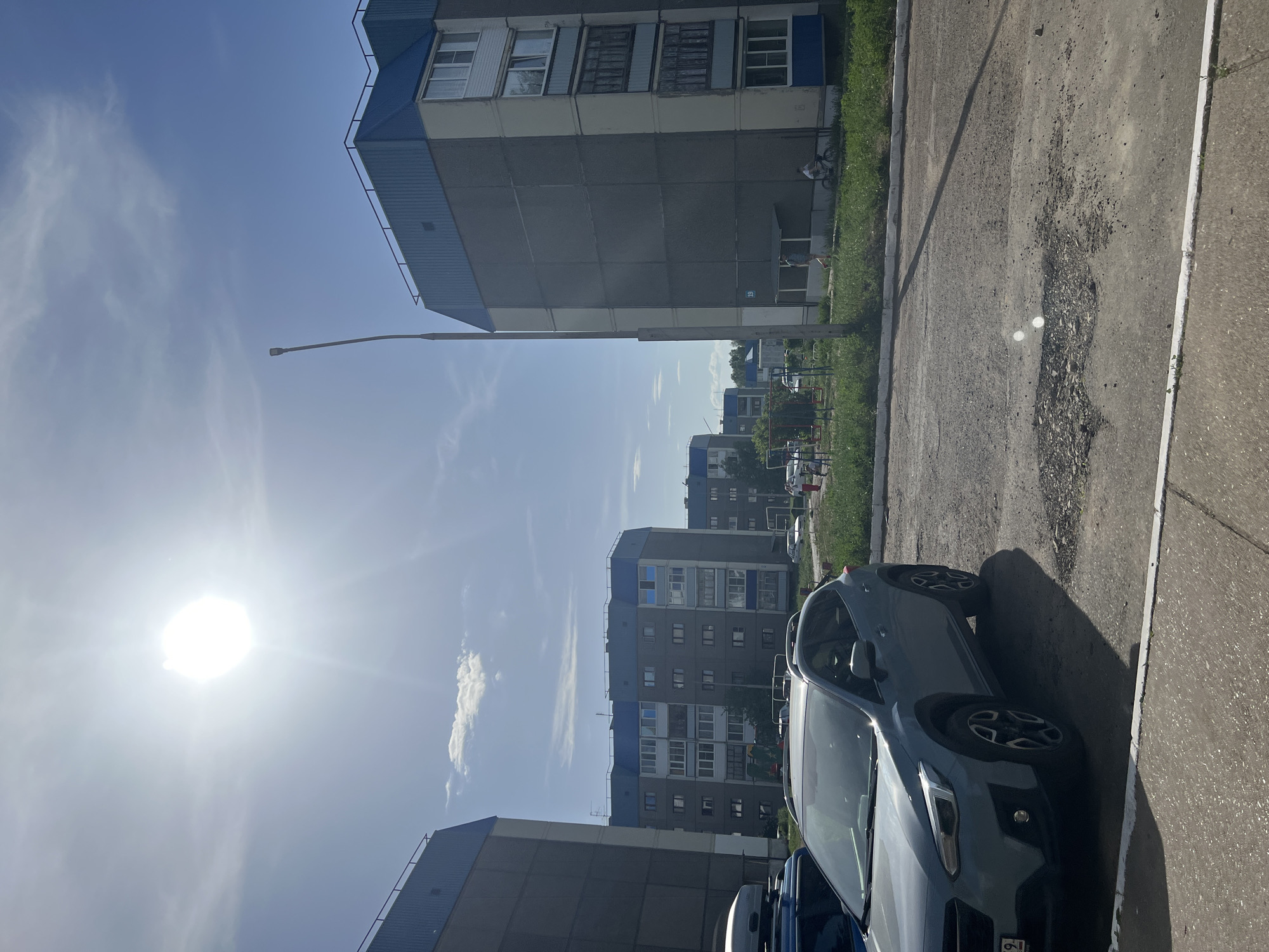 The image depicts a street scene in Марковский, Russia, characterized by Soviet-era apartment buildings under a bright, cloud-streaked sky. A grey Subaru Crosstrek is parked on the right side of the frame along a deteriorating paved road with exposed aggregate. The architecture is typical of post-war Soviet housing, featuring prefabricated concrete blocks with blue roof trim. A tall street light rises in the distance. There are no visible people in the immediate foreground.